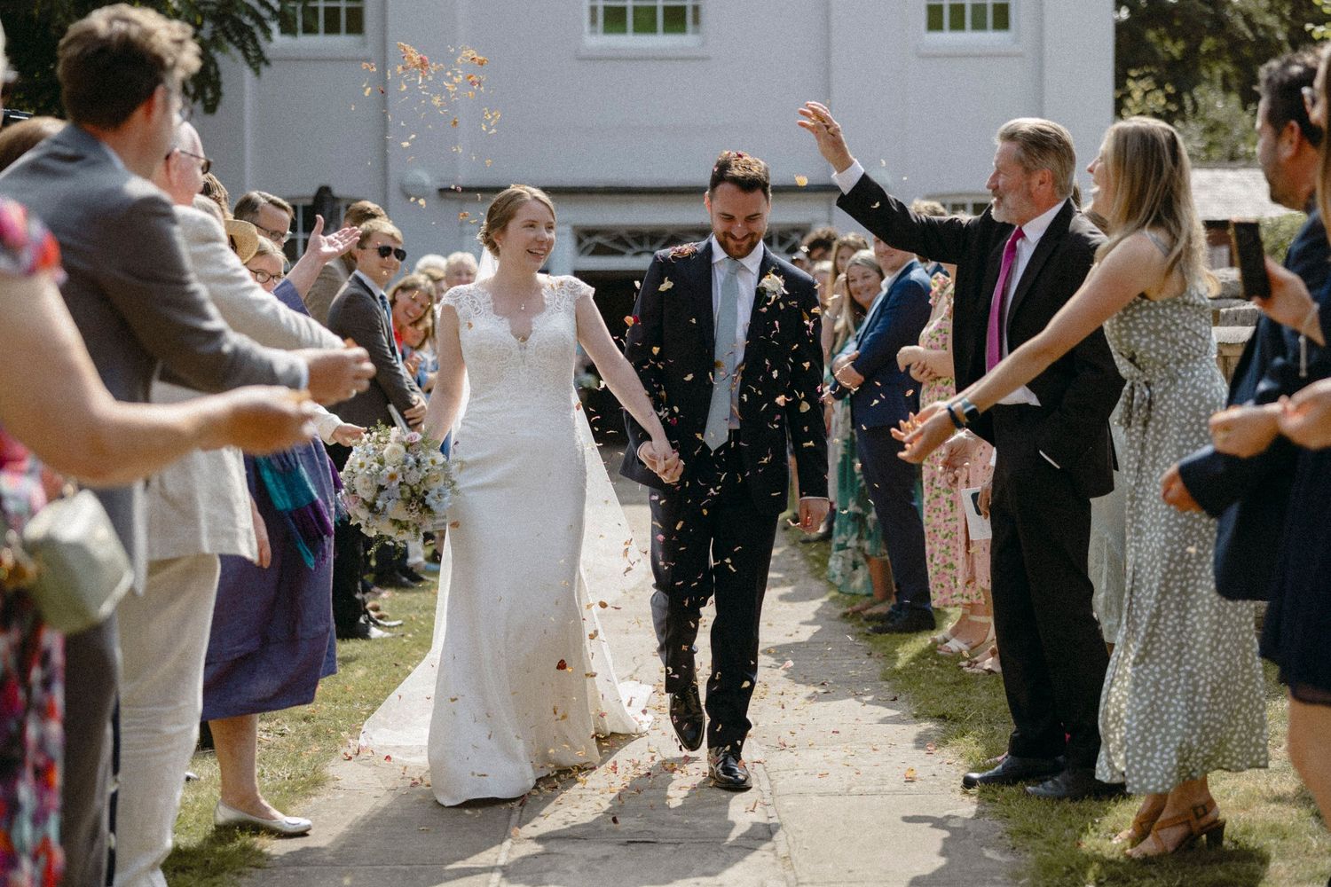 Newlywed couple walks through confetti tunnel surrounded by cheering wedding guests outside a white venue.
