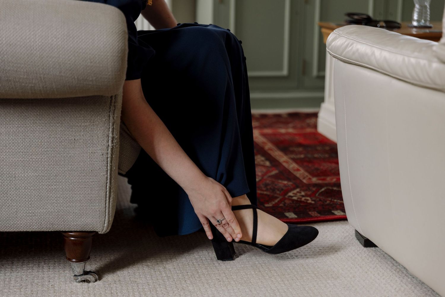 Woman in navy blue dress adjusting black ankle strap heels while seated between two cream upholstered chairs on red rug.