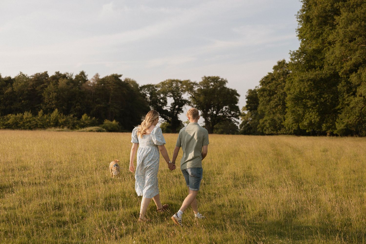 Couple holding hands walking through a sunny green meadow with trees in the background during golden hour.