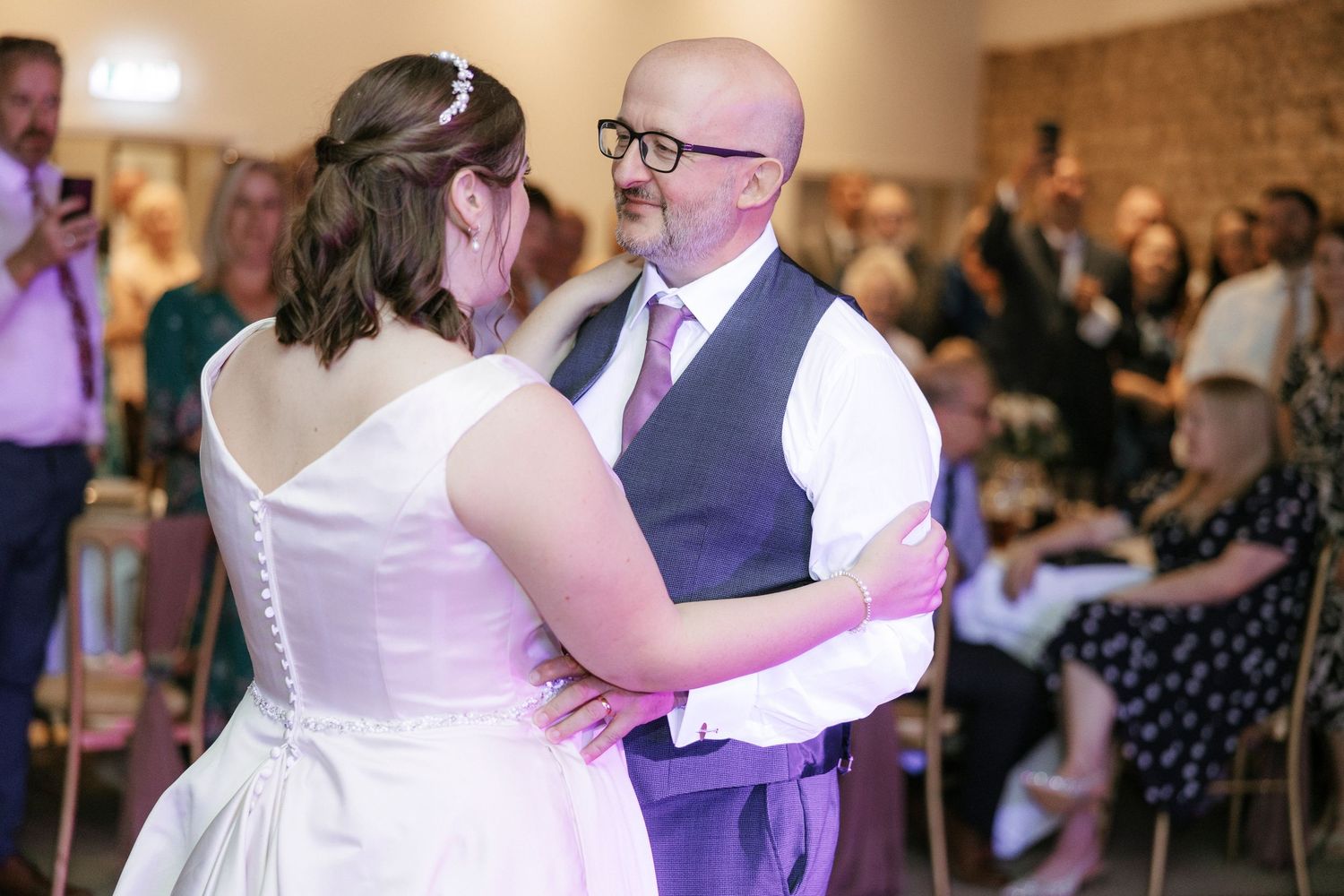 Bride and groom share their first dance at a wedding reception, surrounded by seated guests in a warmly lit venue.