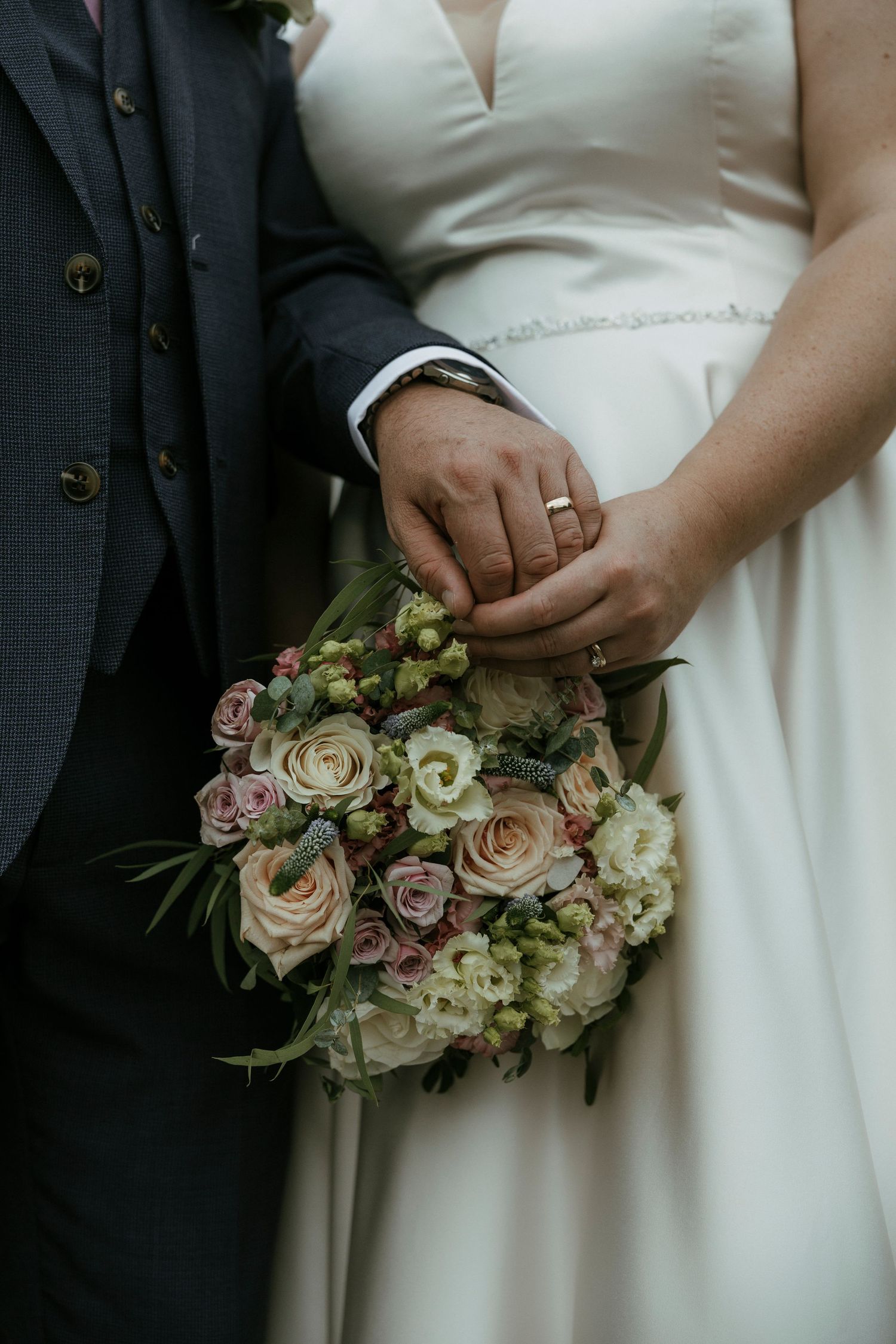 Bride and groom holding a pastel wedding bouquet of roses and greenery, with wedding rings visible on their hands.