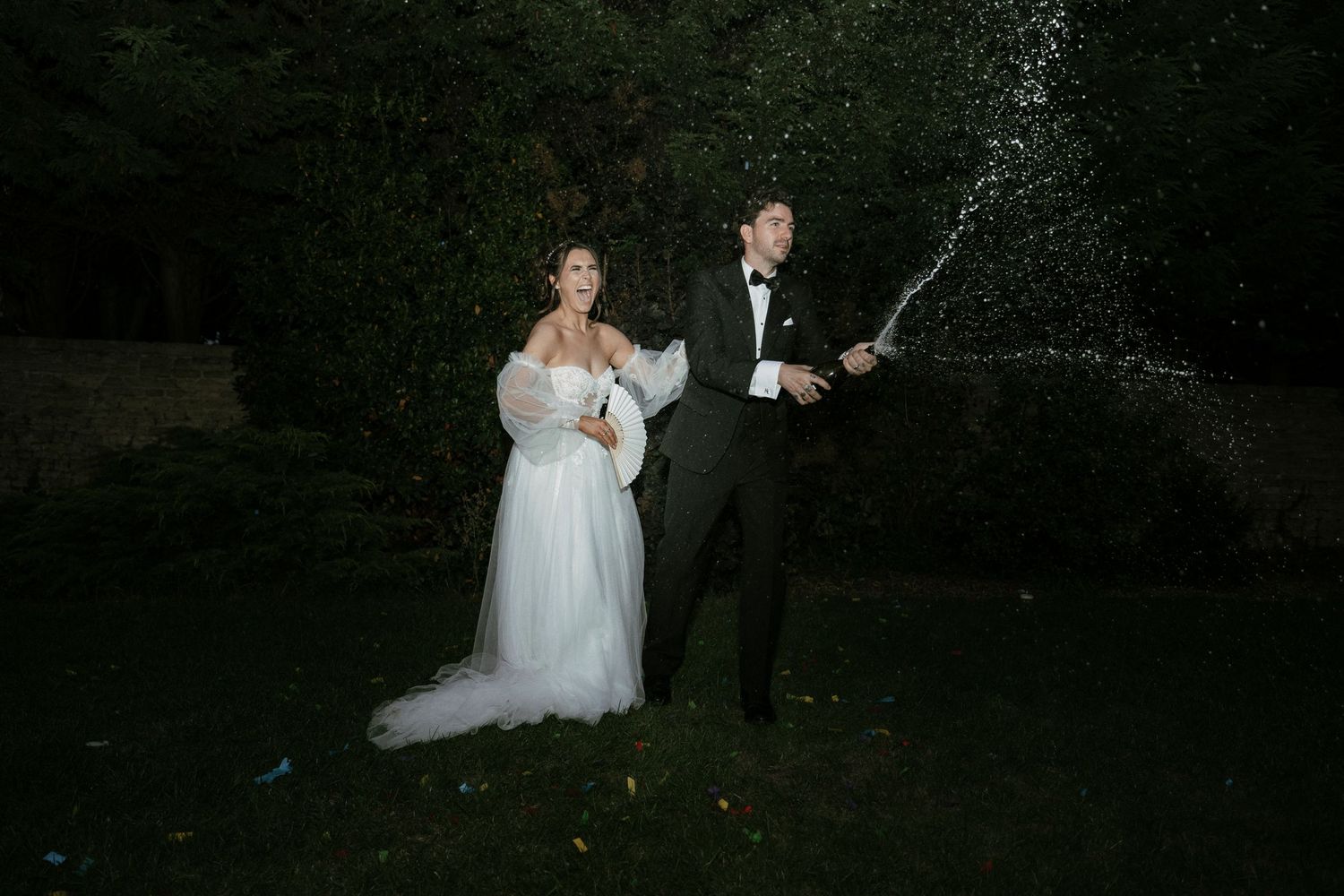 Bride in white gown and groom in tuxedo popping champagne bottle outdoors at night during wedding celebration.