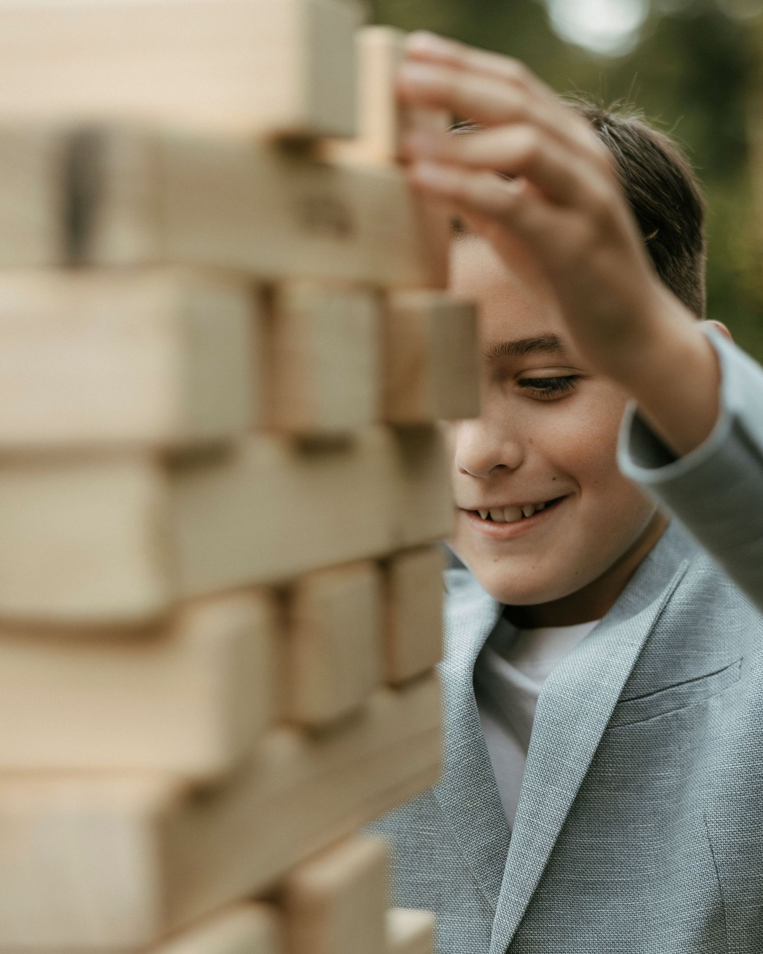 Smiling child in gray blazer playing giant Jenga, carefully removing a wooden block from the tower outdoors.