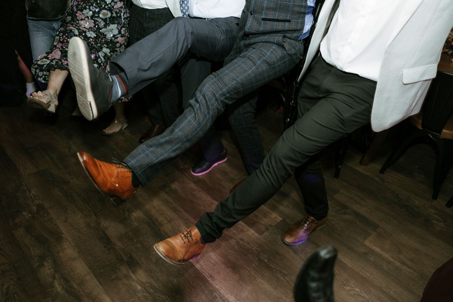 Wedding guests' feet and legs dancing on a dark wooden floor.