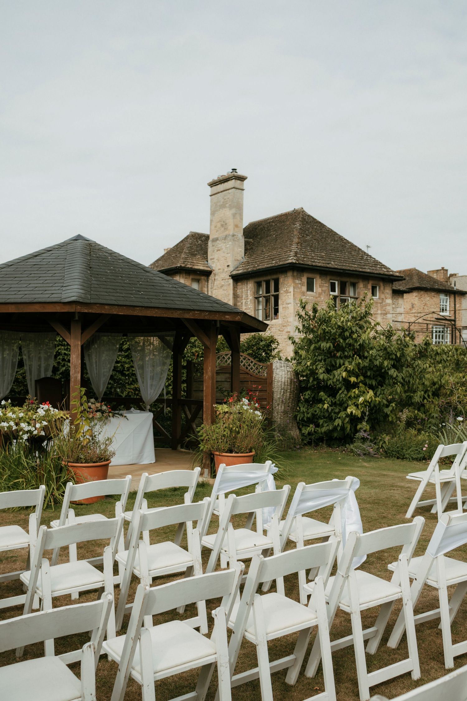 White folding chairs arranged on lawn for outdoor wedding ceremony with wooden gazebo and stone manor house backdrop.