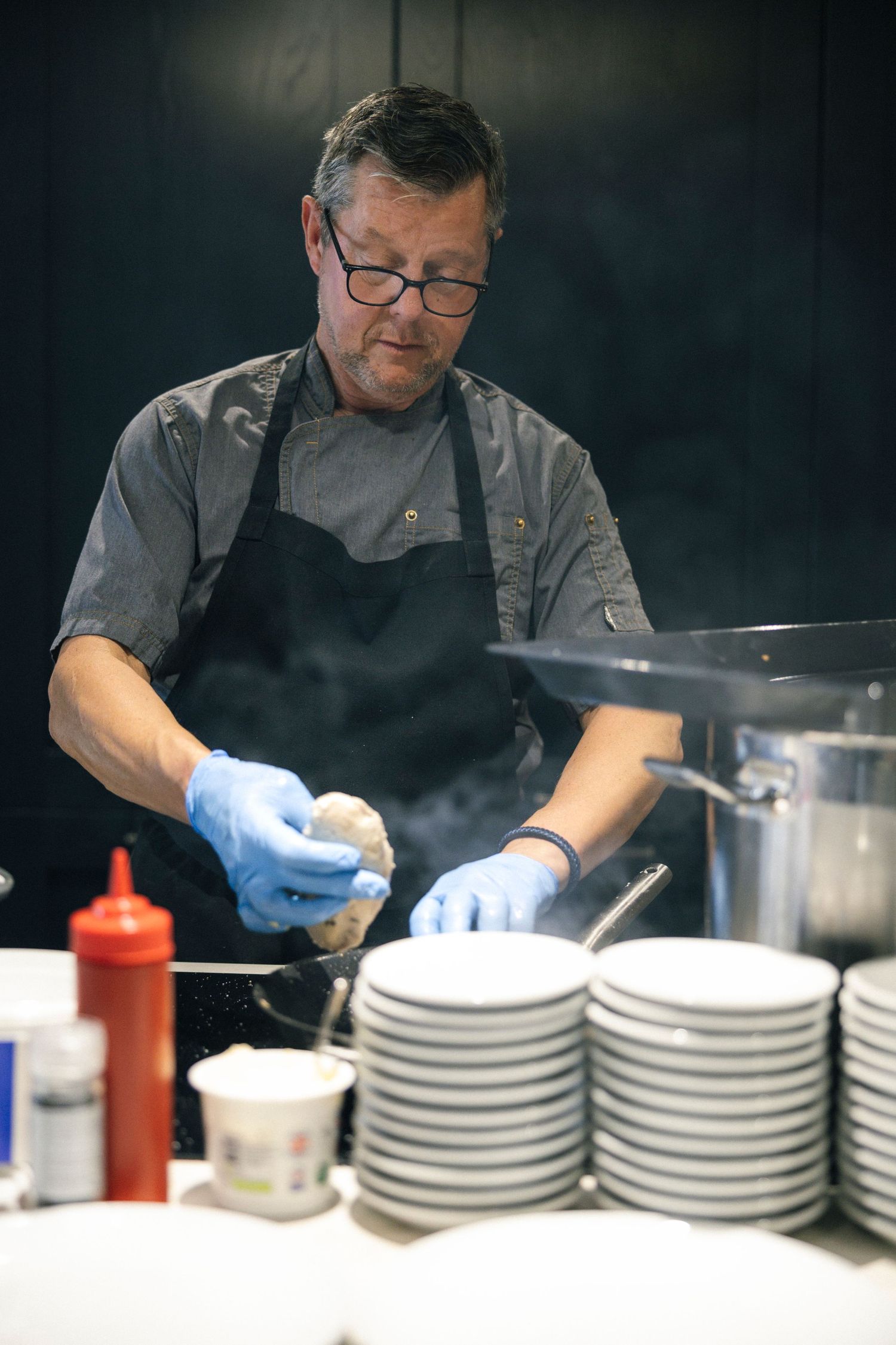 Chef in black apron and blue gloves preparing food beside steaming pot with stacked white plates nearby.