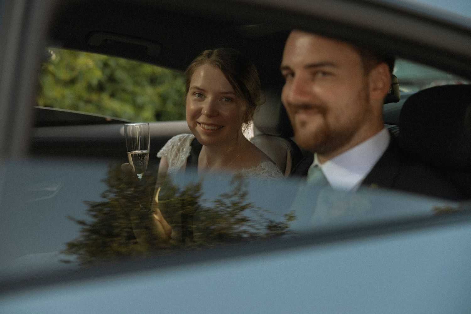 A smiling bride and groom sitting together in a car on their wedding day, with lush green trees visible outside.