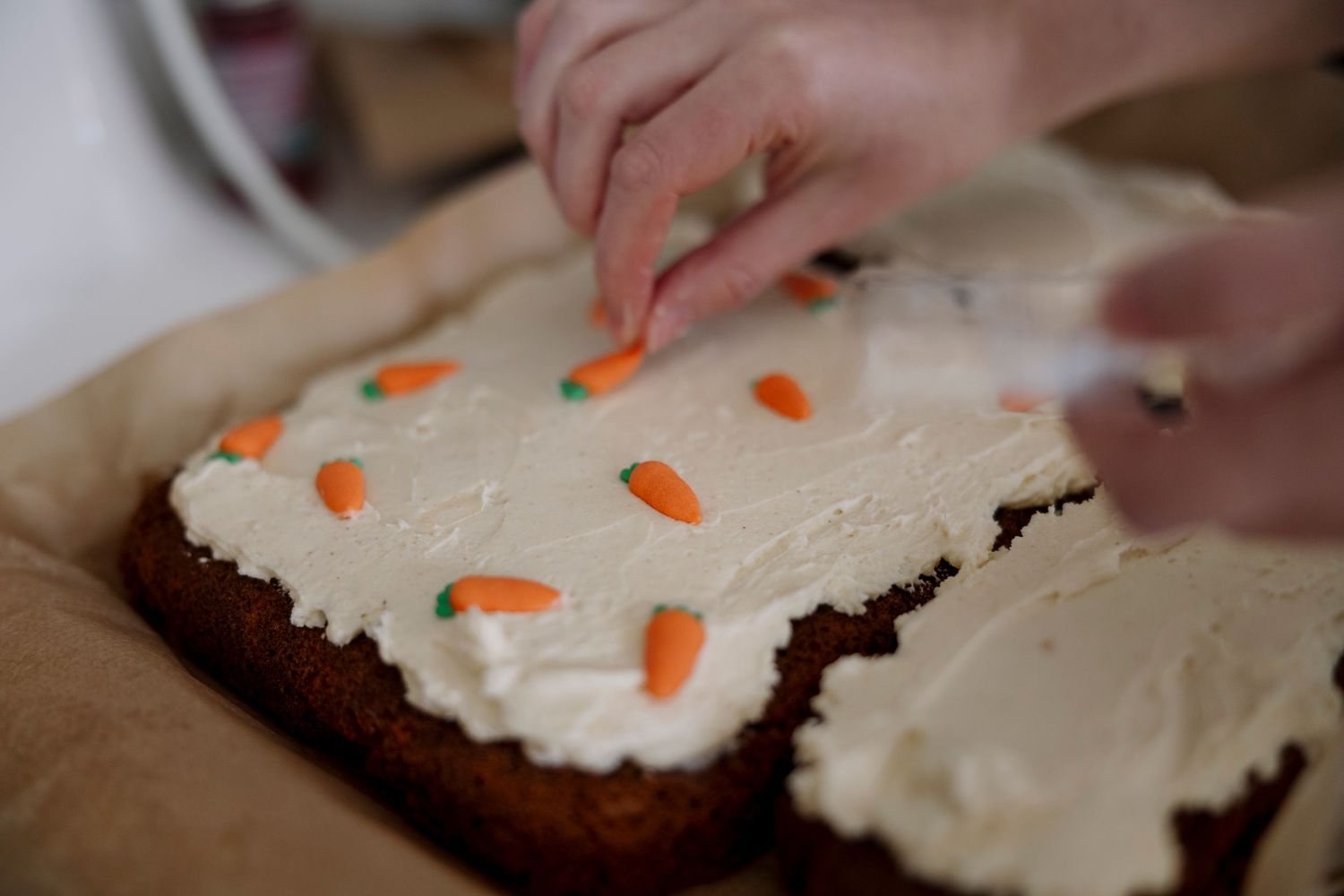 Hands decorating a homemade carrot cake with white cream cheese frosting and small candy carrot sprinkles on top.