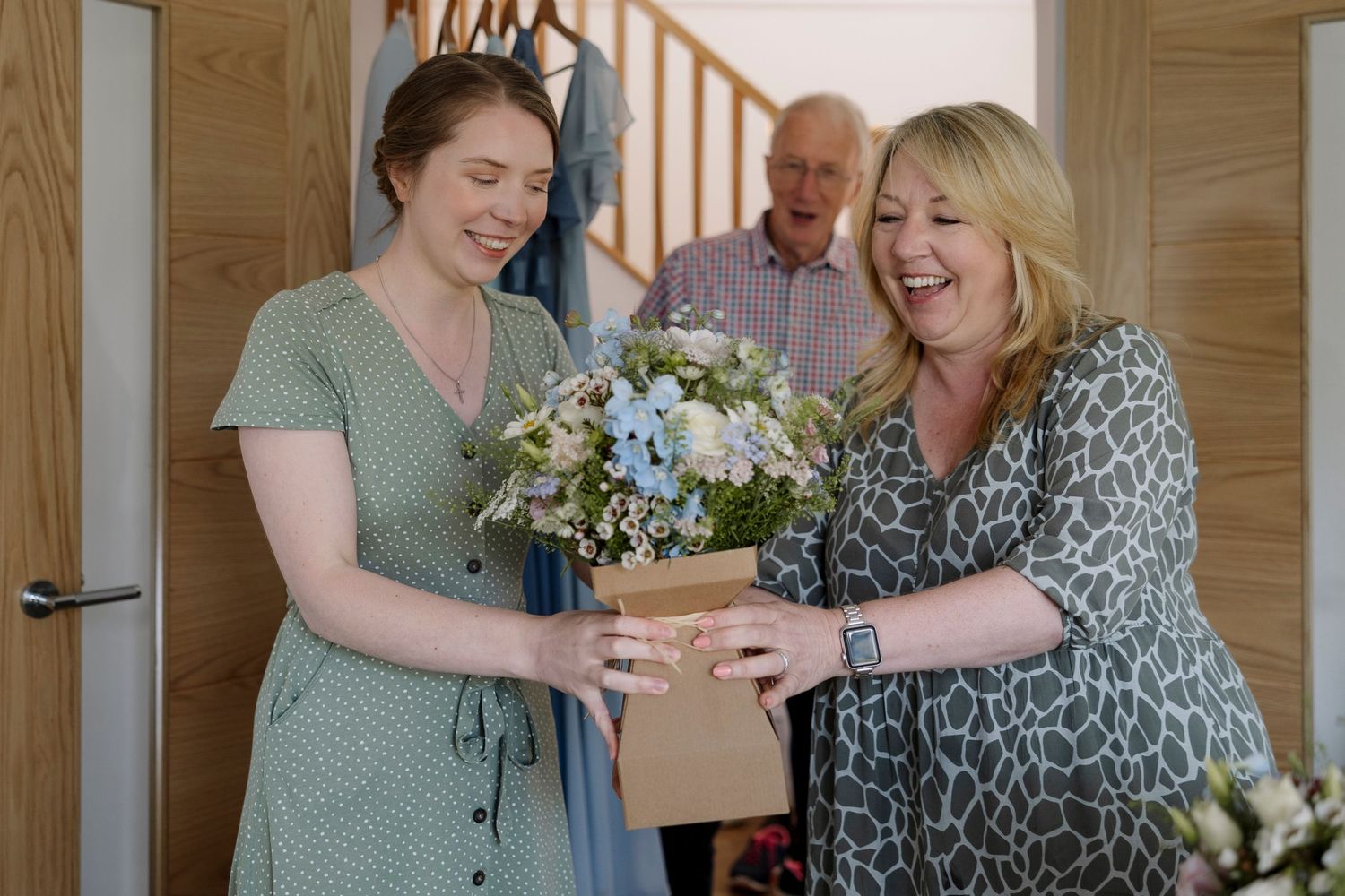 Two women smiling while exchanging a floral bouquet in a box, with an older man watching in the background.