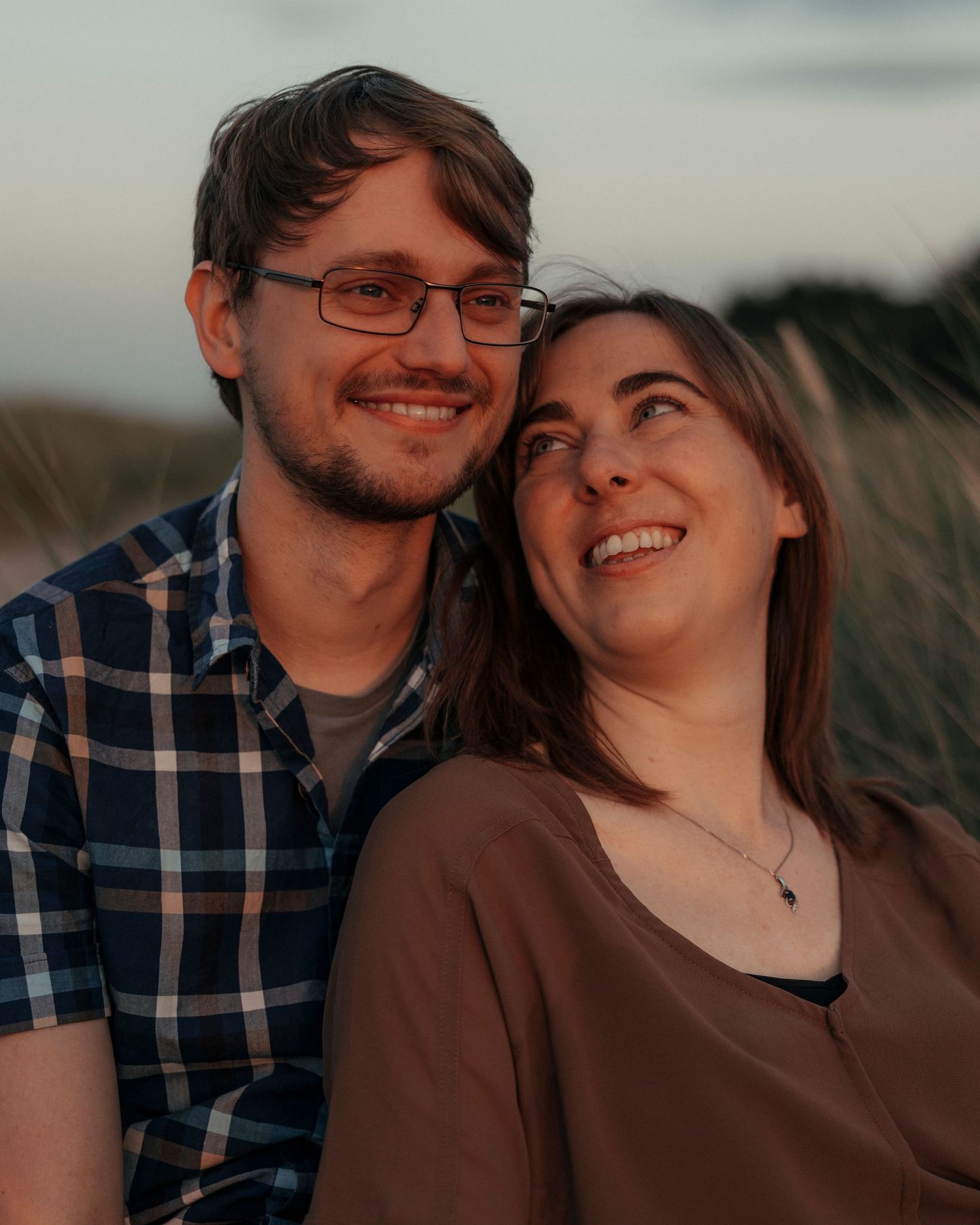 Smiling couple posing closely together outdoors at dusk, man wearing plaid shirt and glasses.
