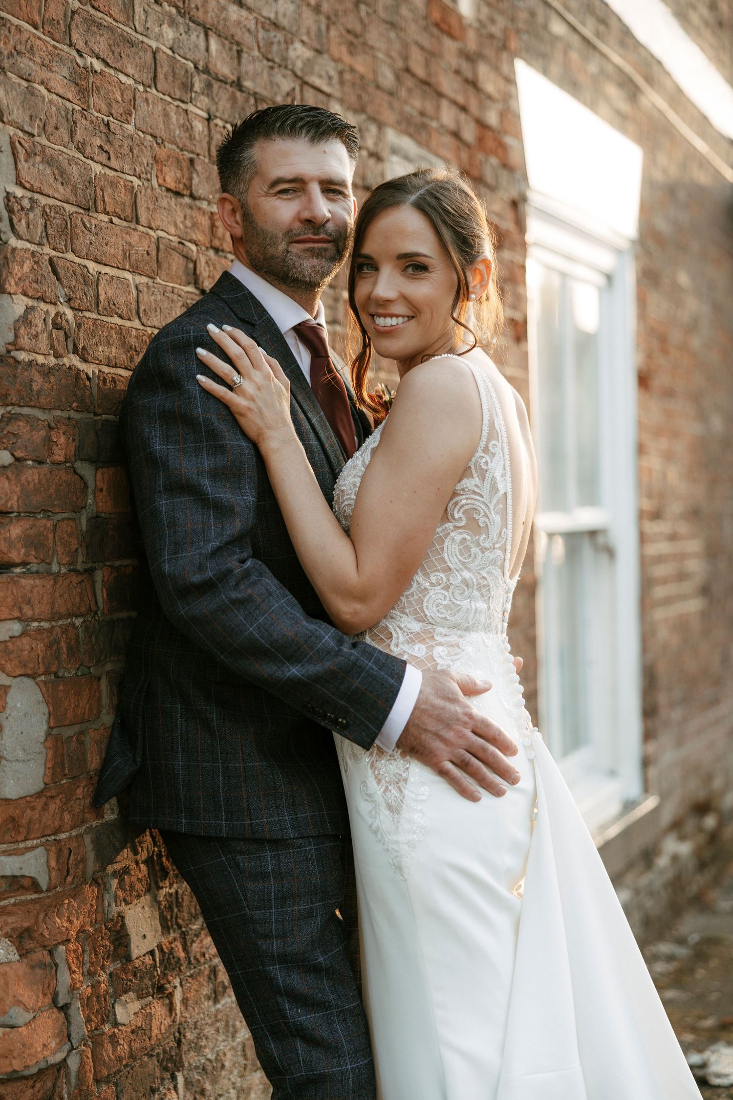 Newlywed couple embracing against rustic brick wall with warm sunlight during sunset wedding photoshoot.