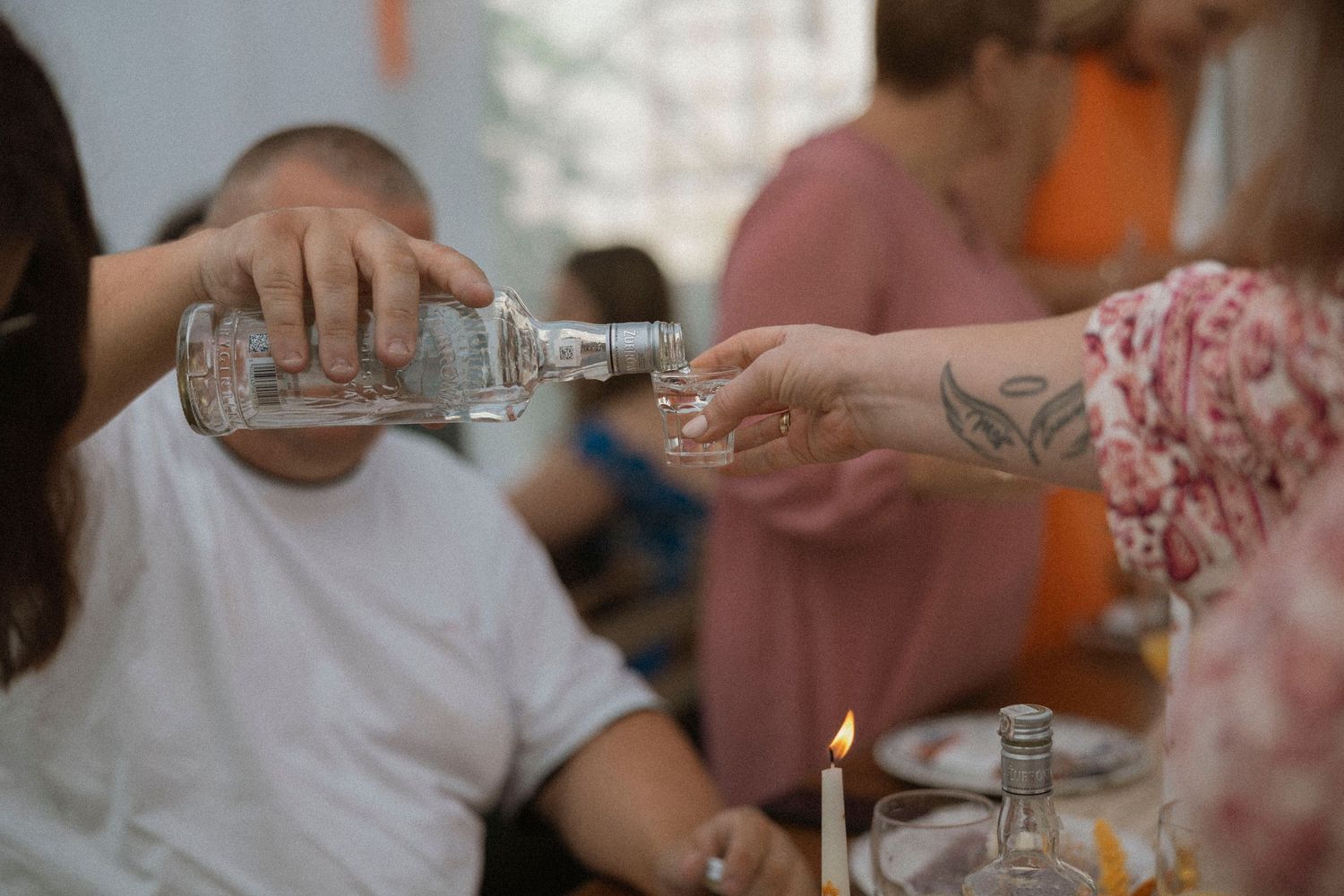 Person pouring clear liquor from a bottle into a shot glass at a social gathering with a lit candle nearby.