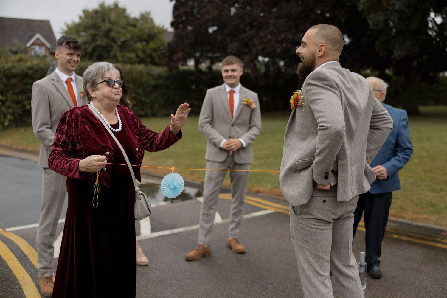 Elderly woman in red velvet dress chats with groomsmen in tan suits at an outdoor wedding gathering.