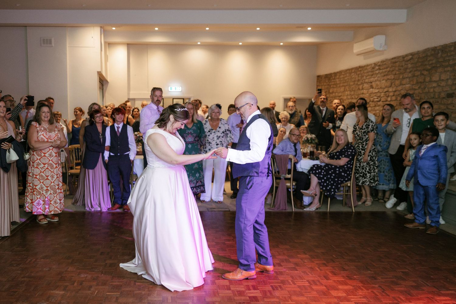 Bride and groom sharing first dance on wooden floor surrounded by wedding guests in bright venue.