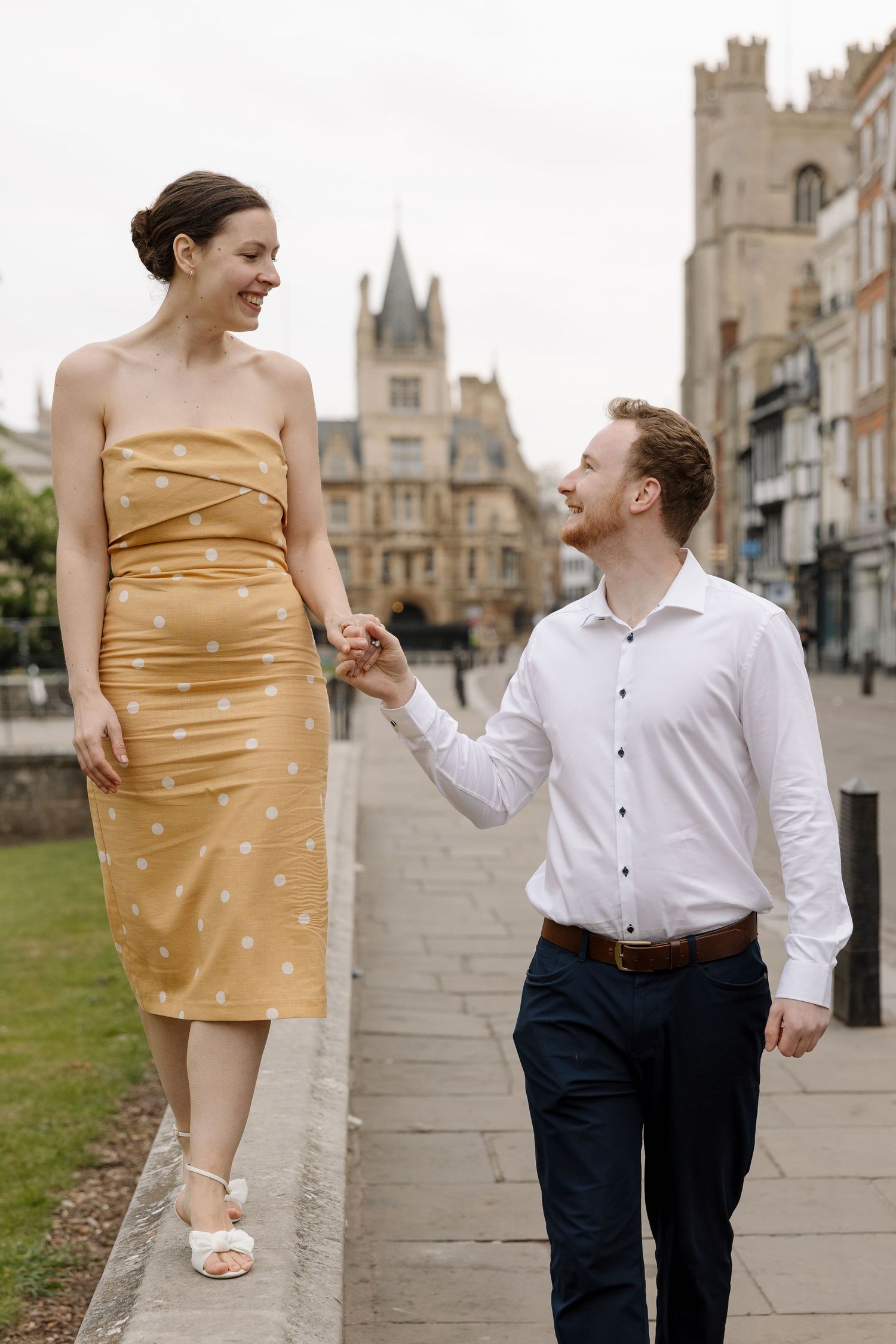 Couple holding hands and smiling at each other while walking along a historic street, woman in gold dress.