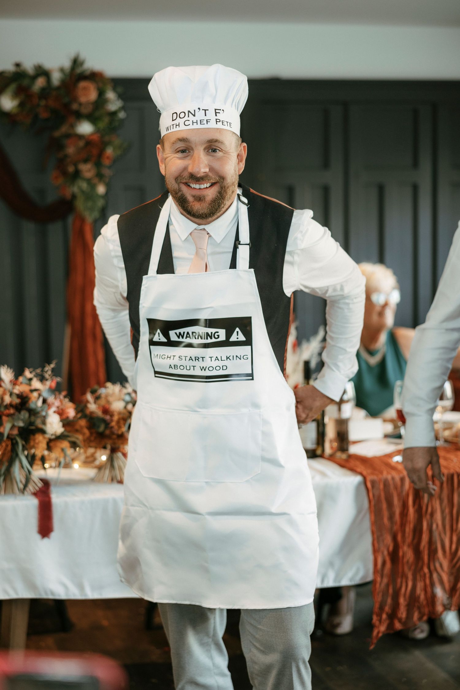 Man in white chef hat and warning apron smiling at a festive indoor event with floral decorations.