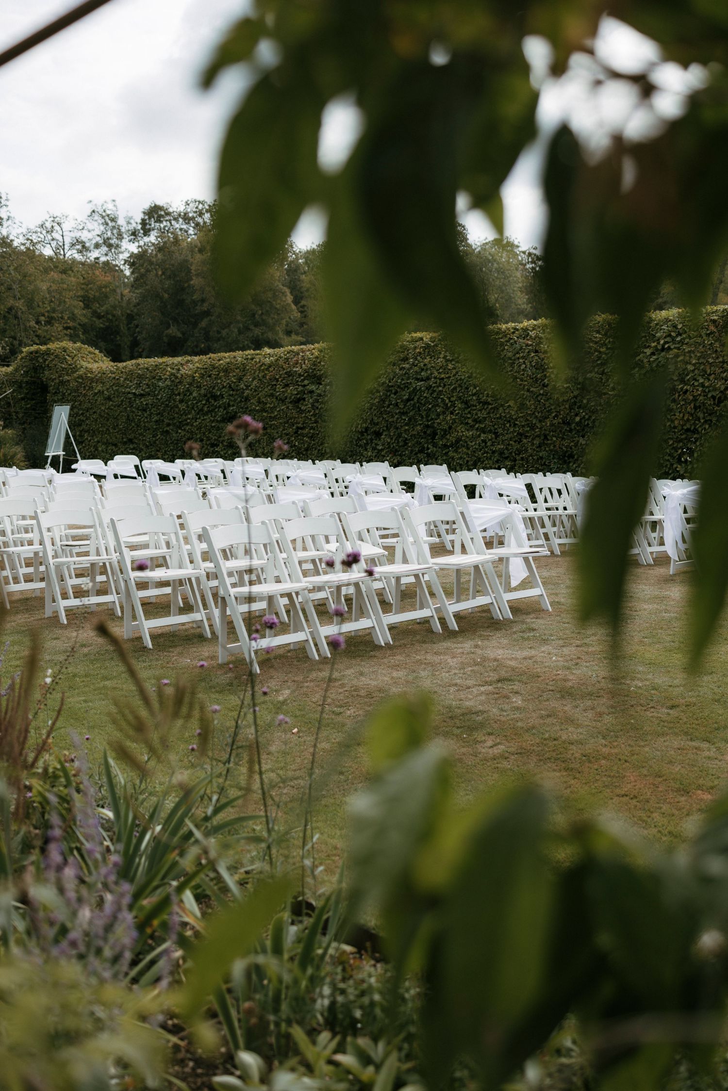 Rows of white folding chairs set up outdoors for a garden wedding ceremony, surrounded by lush green hedges.