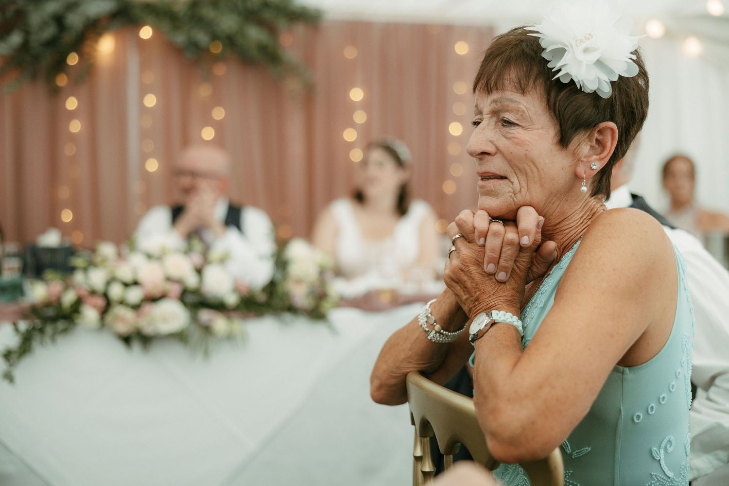 Elderly woman in teal dress with floral hair accessory clasping hands at a wedding reception.