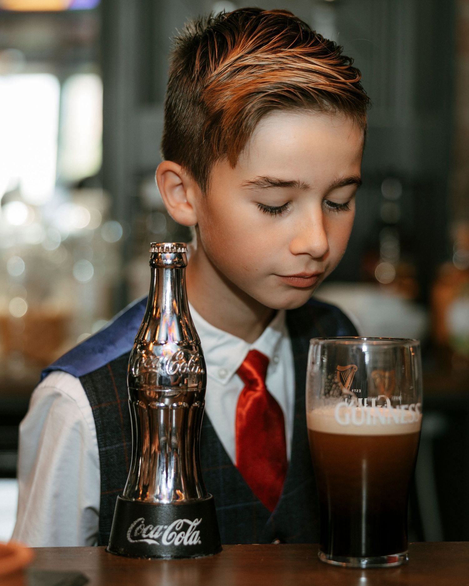 Young boy in suit with red tie sits at bar beside Coca-Cola bottle and Guinness glass.