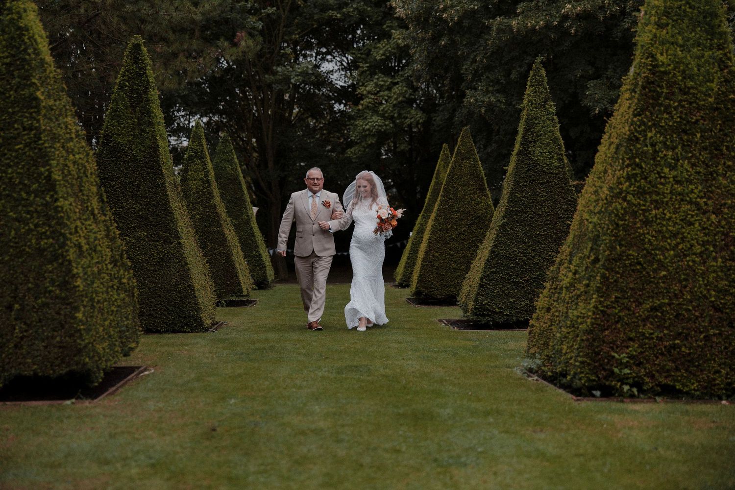 Bride and groom walking hand in hand through a formal garden with tall pyramid-shaped topiary hedges on their wedding day.
