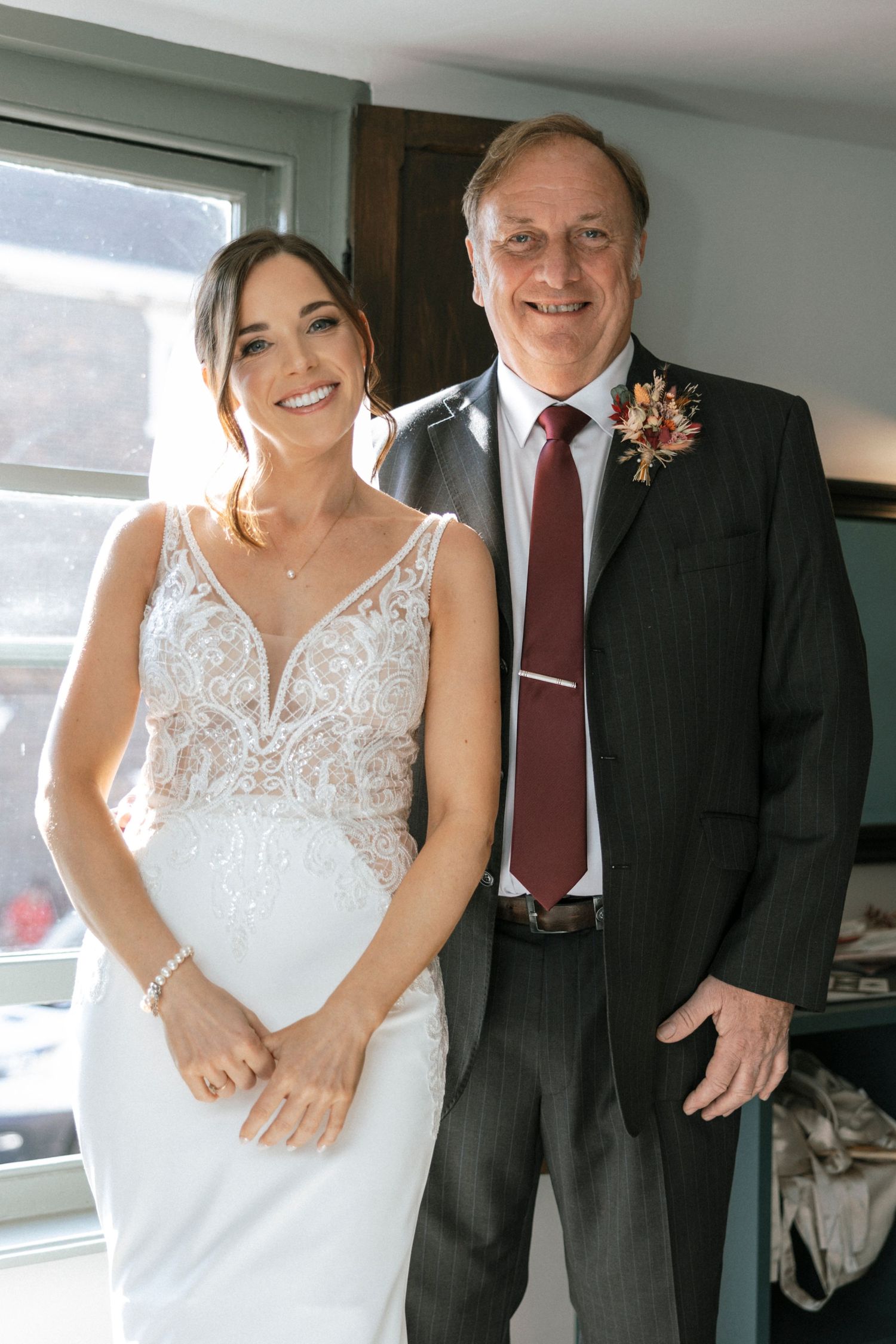 A wedding portrait shows a bride wearing an elegant lace gown standing next to someone in a dark suit with a boutonniere.