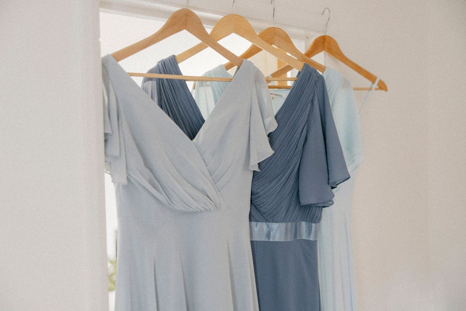 Three mismatched dusty blue bridesmaid dresses hanging on wooden hangers against a white wall.