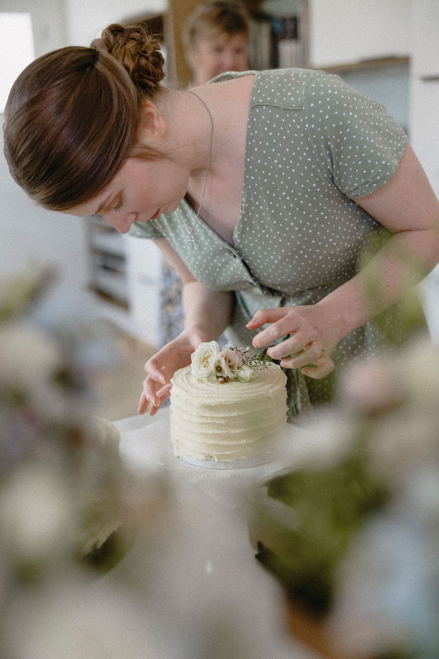 Woman in green polka dot dress decorating a white layered cake with floral toppings in a bright kitchen.