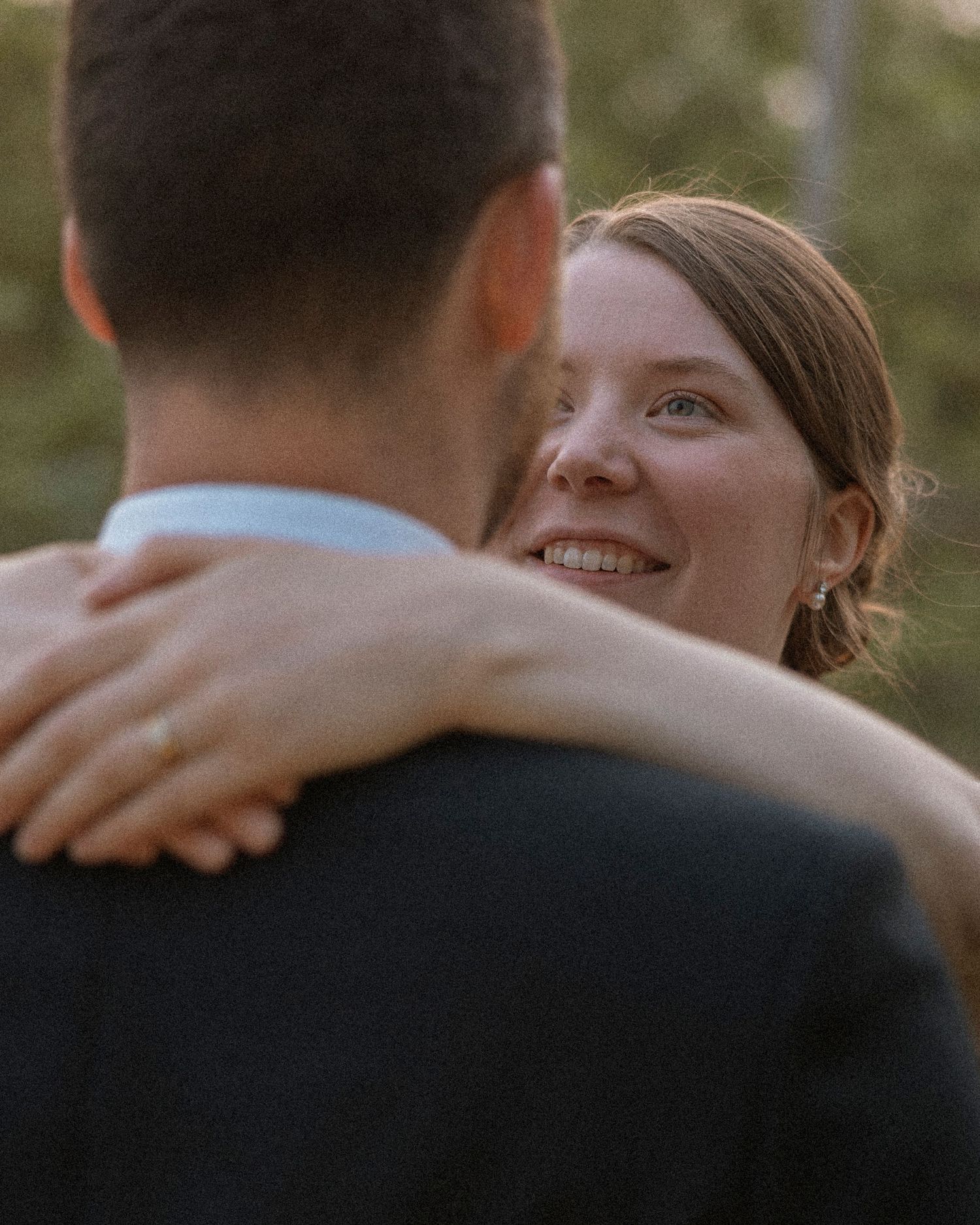 A smiling bride with her arms around a groom in a dark suit, sharing a romantic dance outdoors at golden hour.