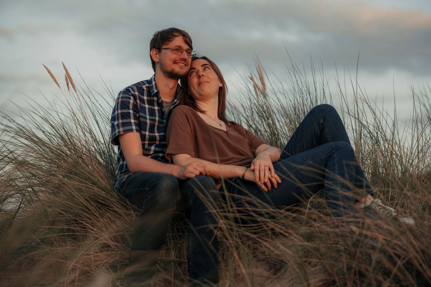 Couple sitting together peacefully in tall grass under a cloudy sky.