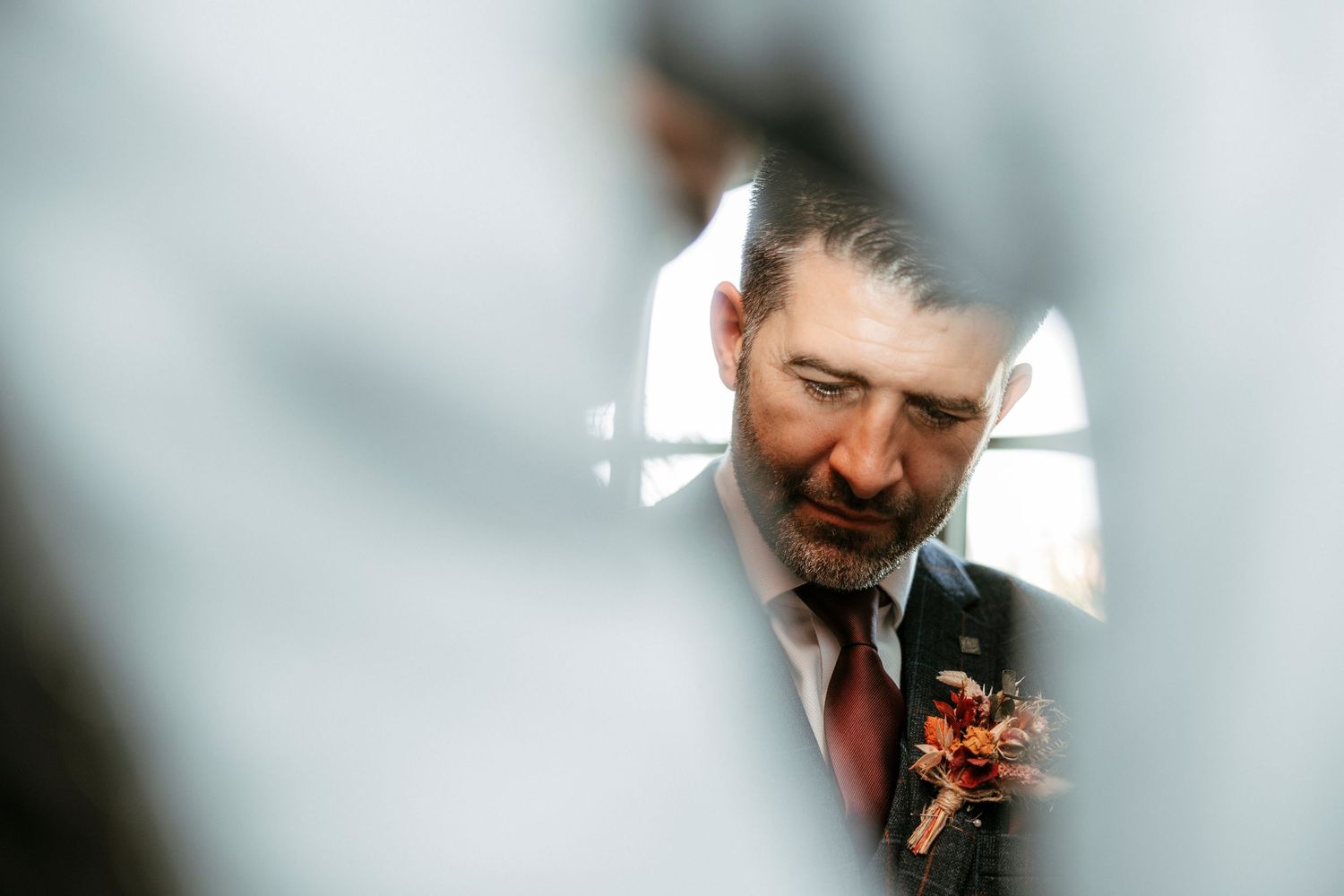 Groom in suit with floral boutonniere seen through gap, candid wedding portrait with soft bokeh background.