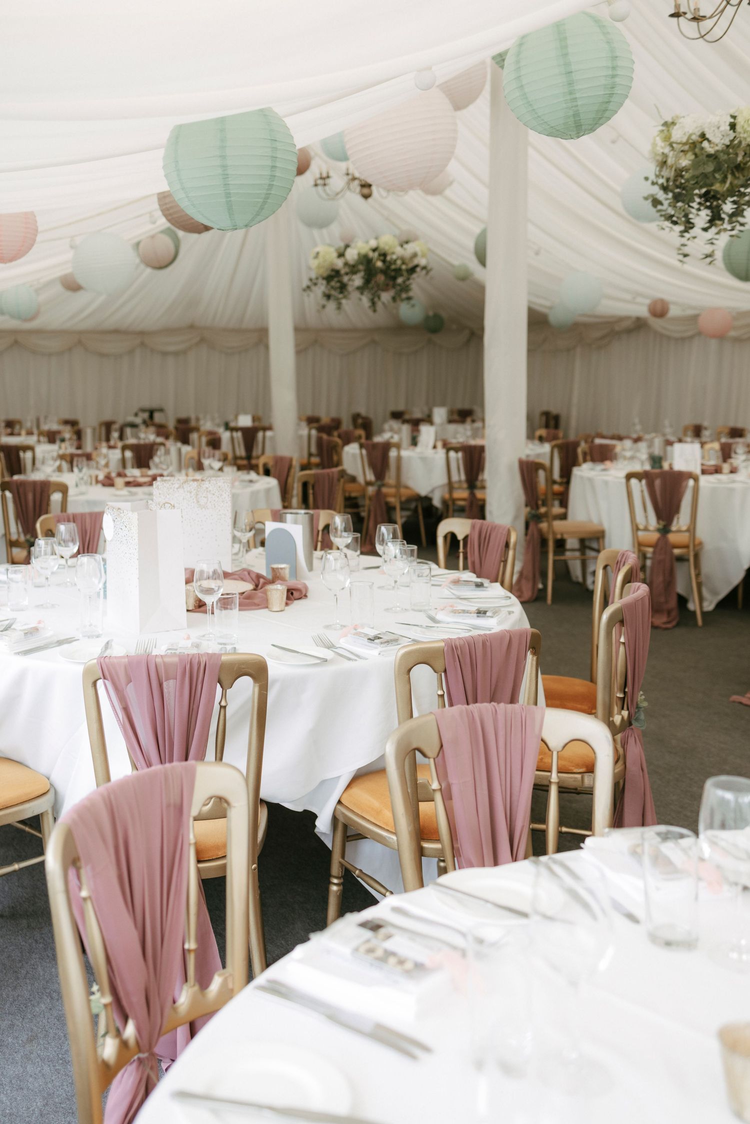 Elegant wedding marquee with round tables, dusty rose chair sashes, gold chairs, and pastel paper lanterns hanging from draped ceiling.