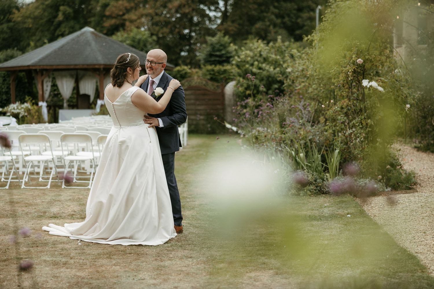 Bride and groom share first dance in lush garden wedding venue with white chairs and floral arrangements.
