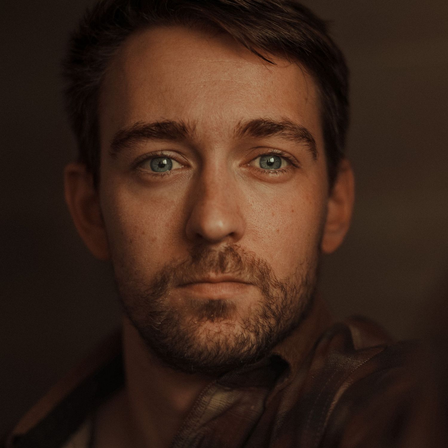 Close-up portrait of a young man with green eyes, light stubble, and freckles against a dark background.