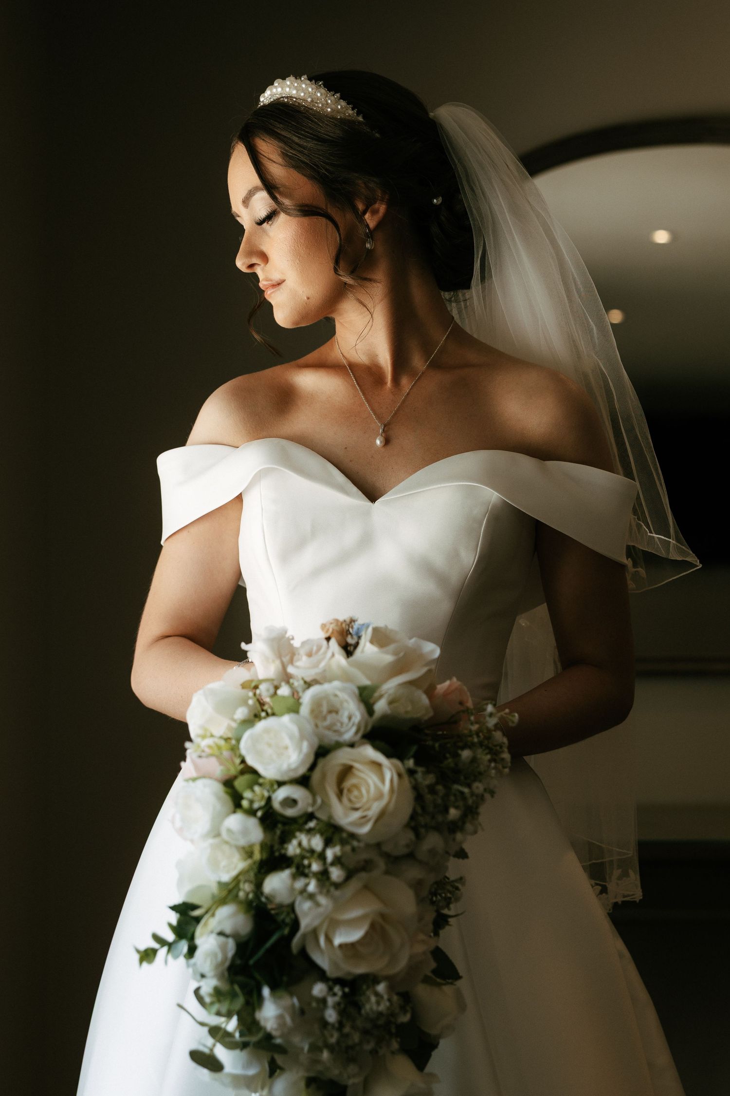 Elegant bride in off-shoulder white gown holding bouquet of white roses, wearing tiara and veil indoors.