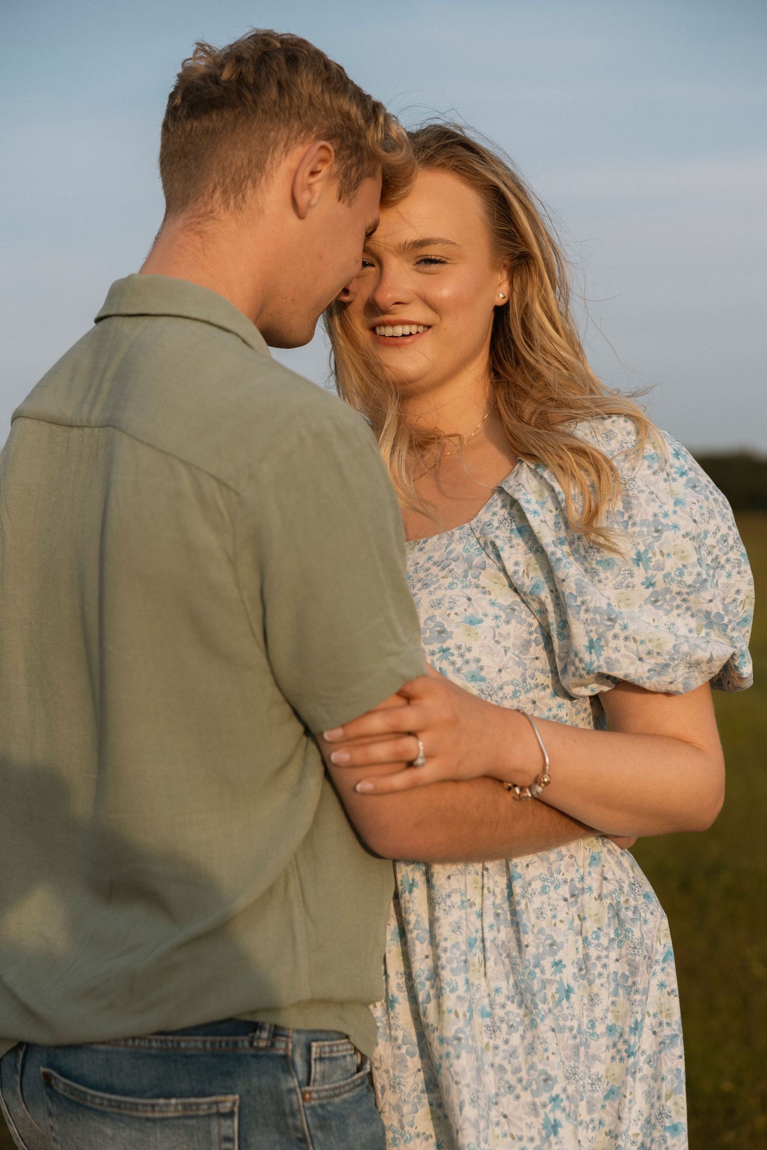 A smiling blonde woman in a floral dress embraces a man in a green shirt in a sunlit field.