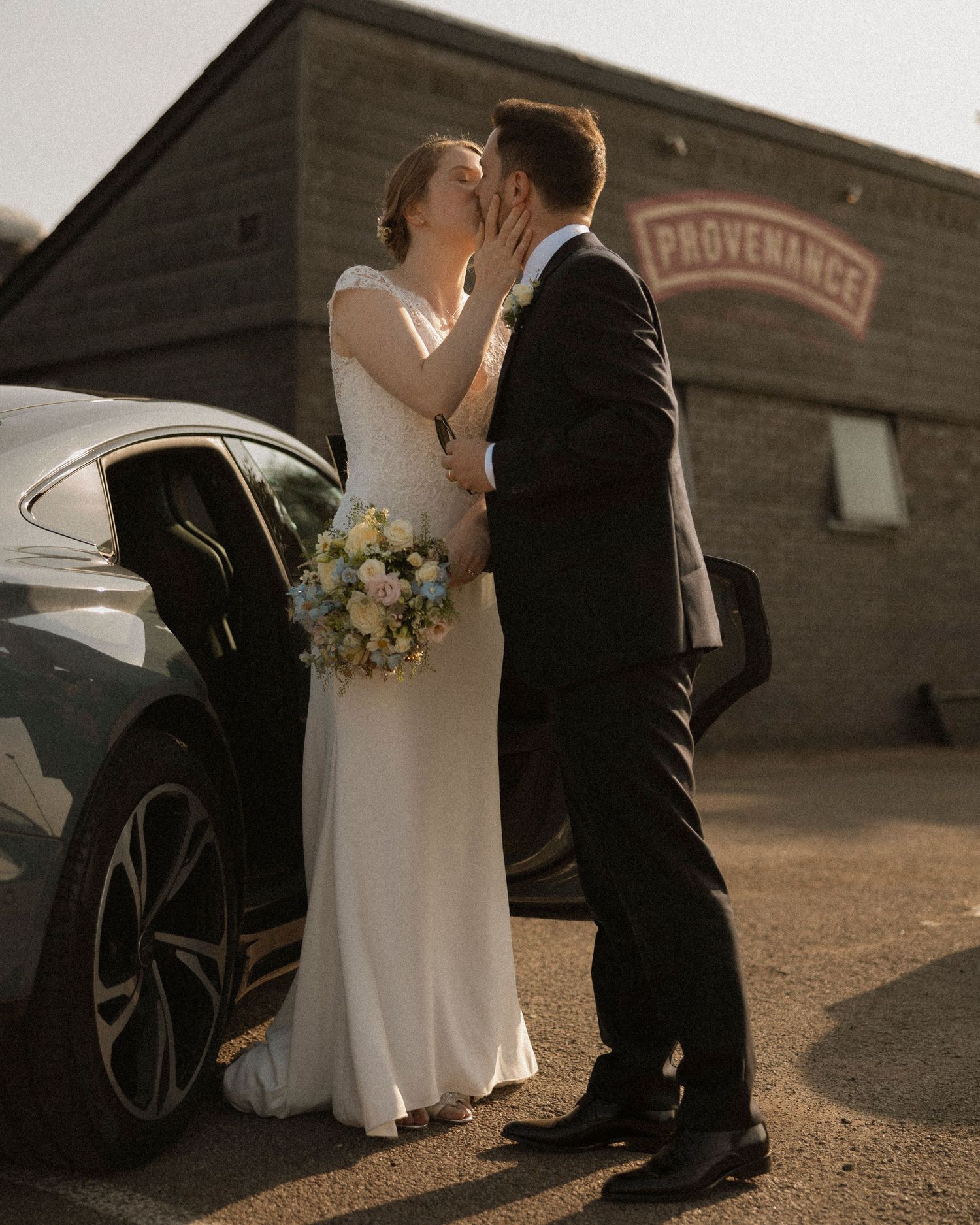 A bride and groom share a kiss outside the Provenance venue beside a black car, bride holding a pastel bouquet.