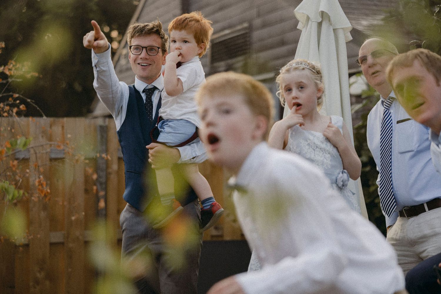Children and adults laughing and pointing outdoors at a wedding celebration, dressed in formal attire.