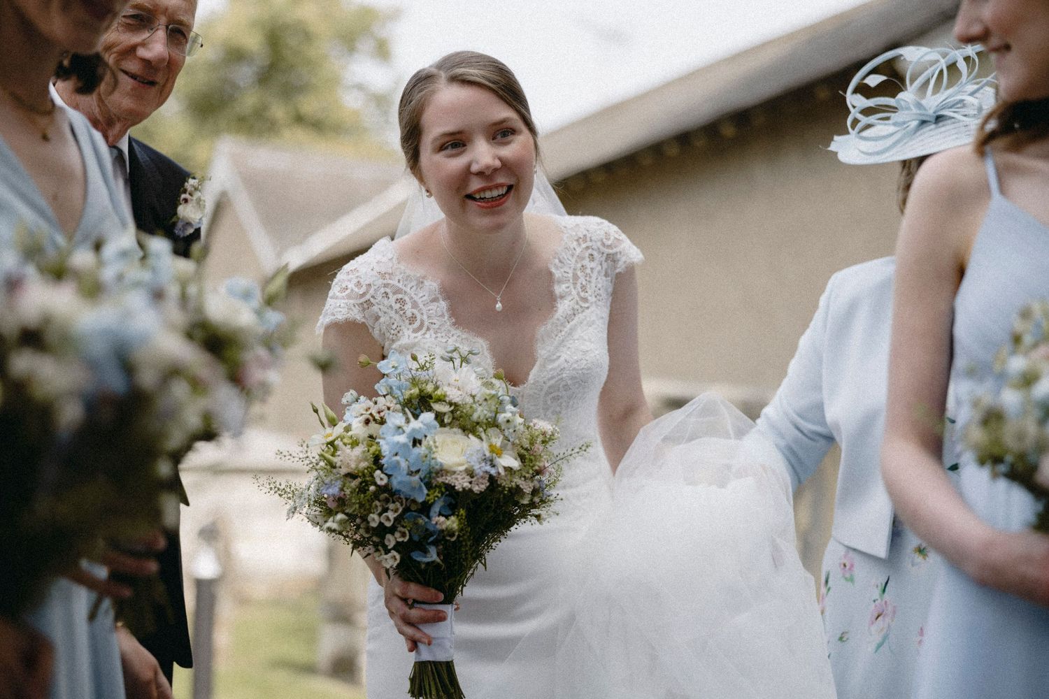 Smiling bride in white lace dress holding blue and white floral bouquet surrounded by guests at outdoor wedding ceremony.