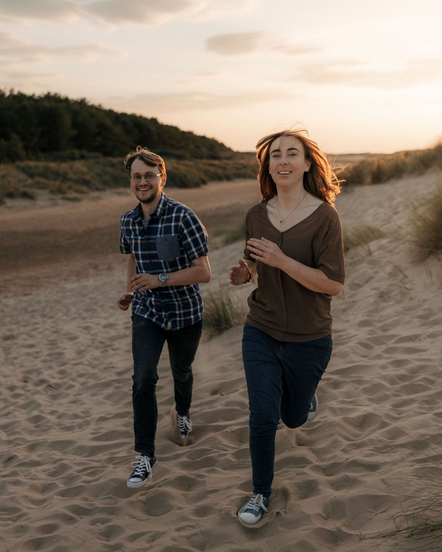 Smiling couple running along sandy beach dunes at golden sunset.