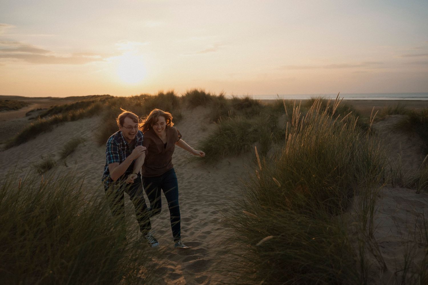 Couple running playfully through beach grass at sunset.