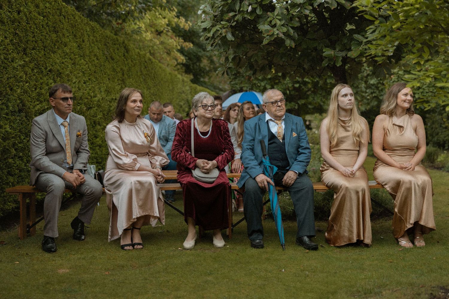 Wedding guests seated outdoors on wooden benches in a lush garden, dressed in elegant formal attire.