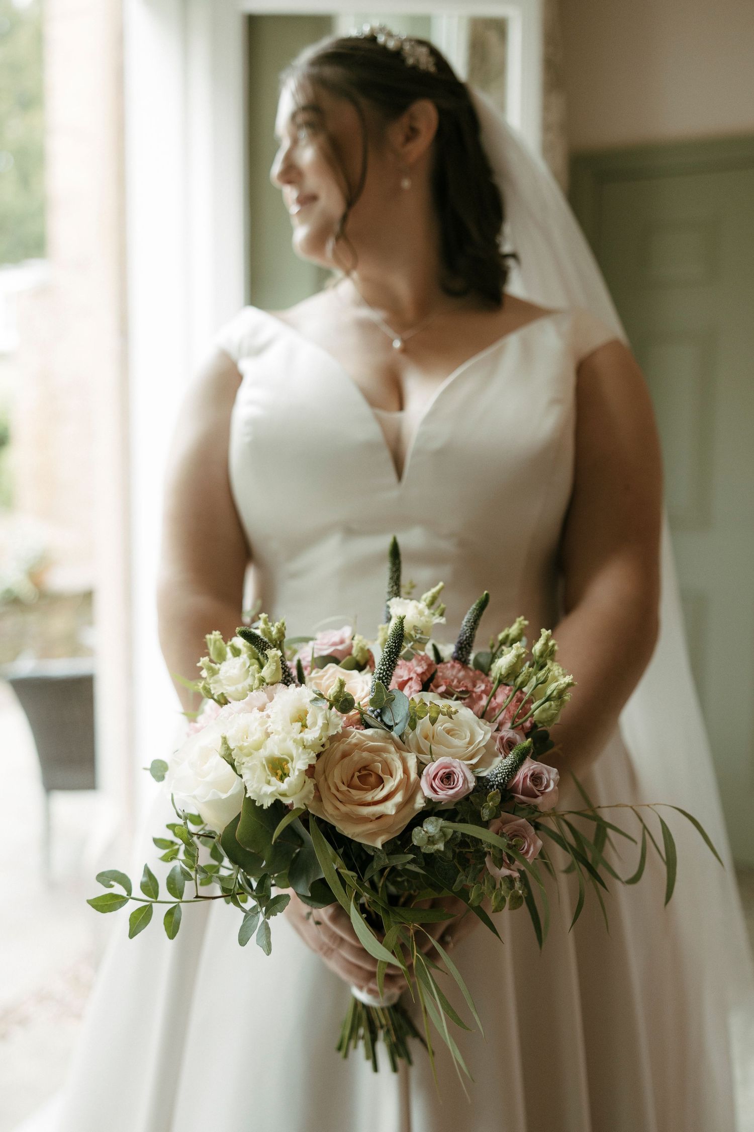 Bride in white gown holding elegant bouquet of roses and greenery by window light.