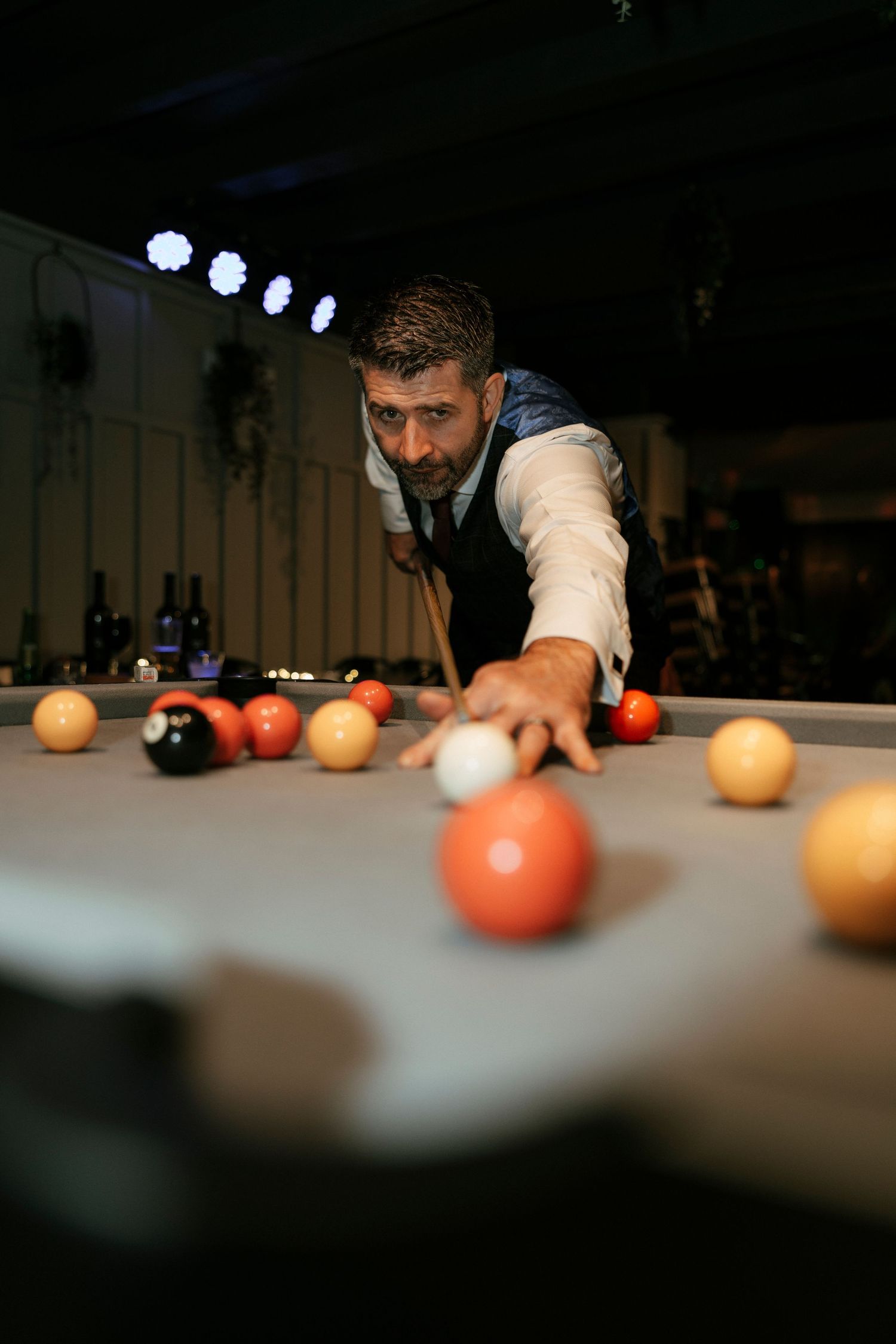 Person in vest leans over pool table preparing to take a shot in dimly lit billiards room.