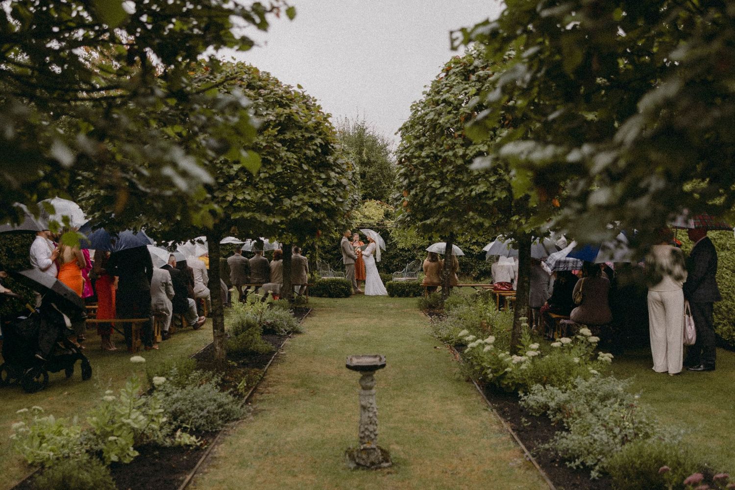 Outdoor garden wedding ceremony with guests lining a lush green aisle leading to a floral arch.