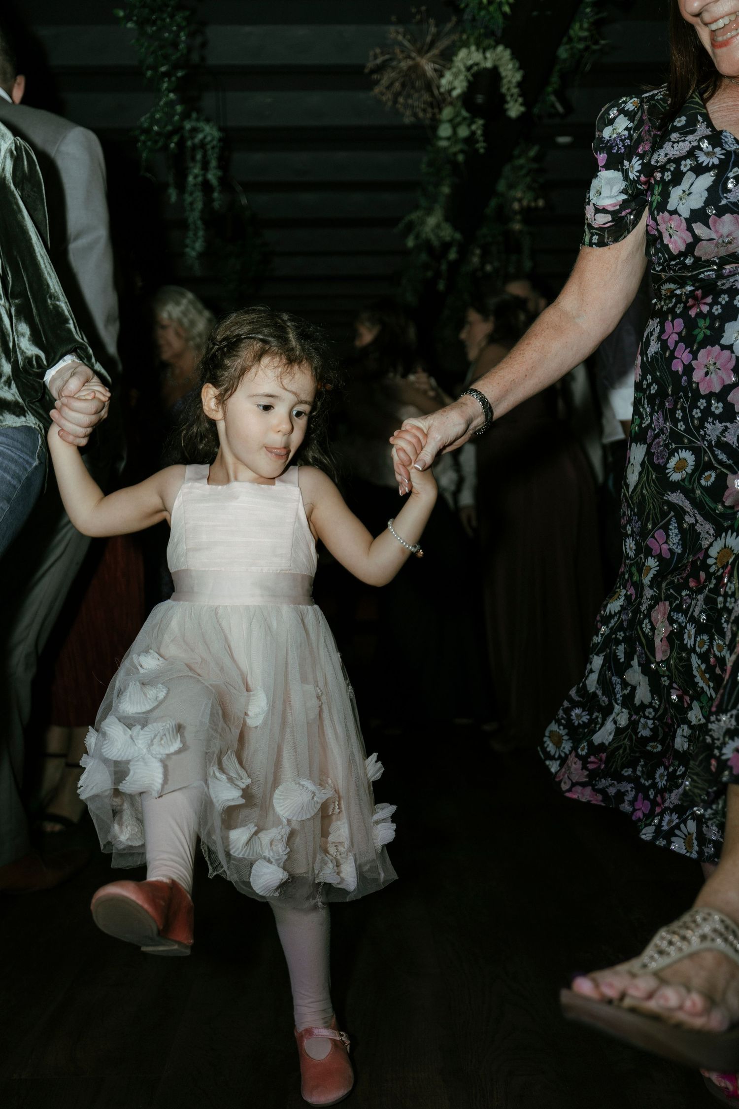 Young flower girl in white tulle dress dancing joyfully at a wedding reception, holding hands with guests.