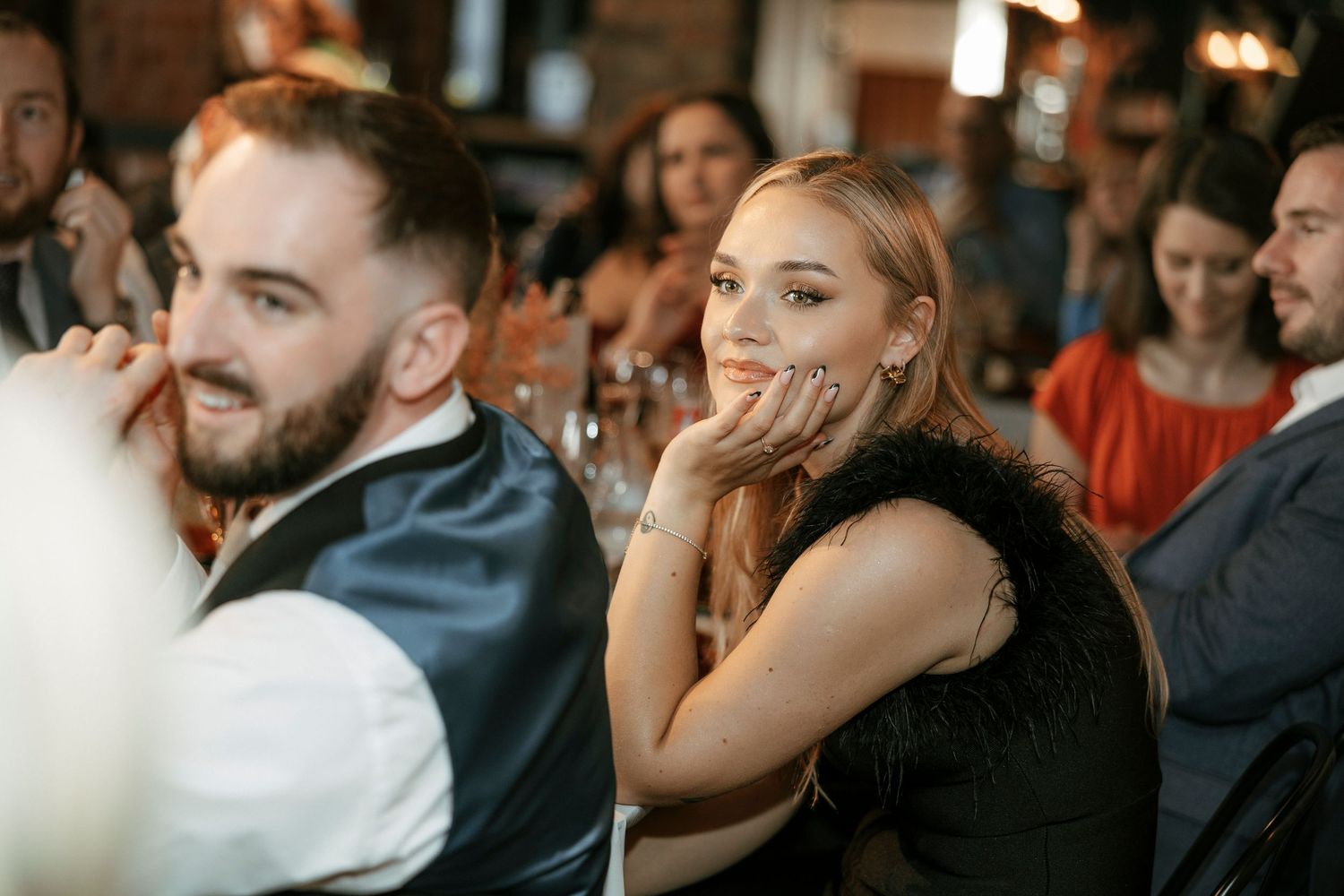 A smiling young woman in a black dress sits beside a bearded man at a lively indoor dinner event.
