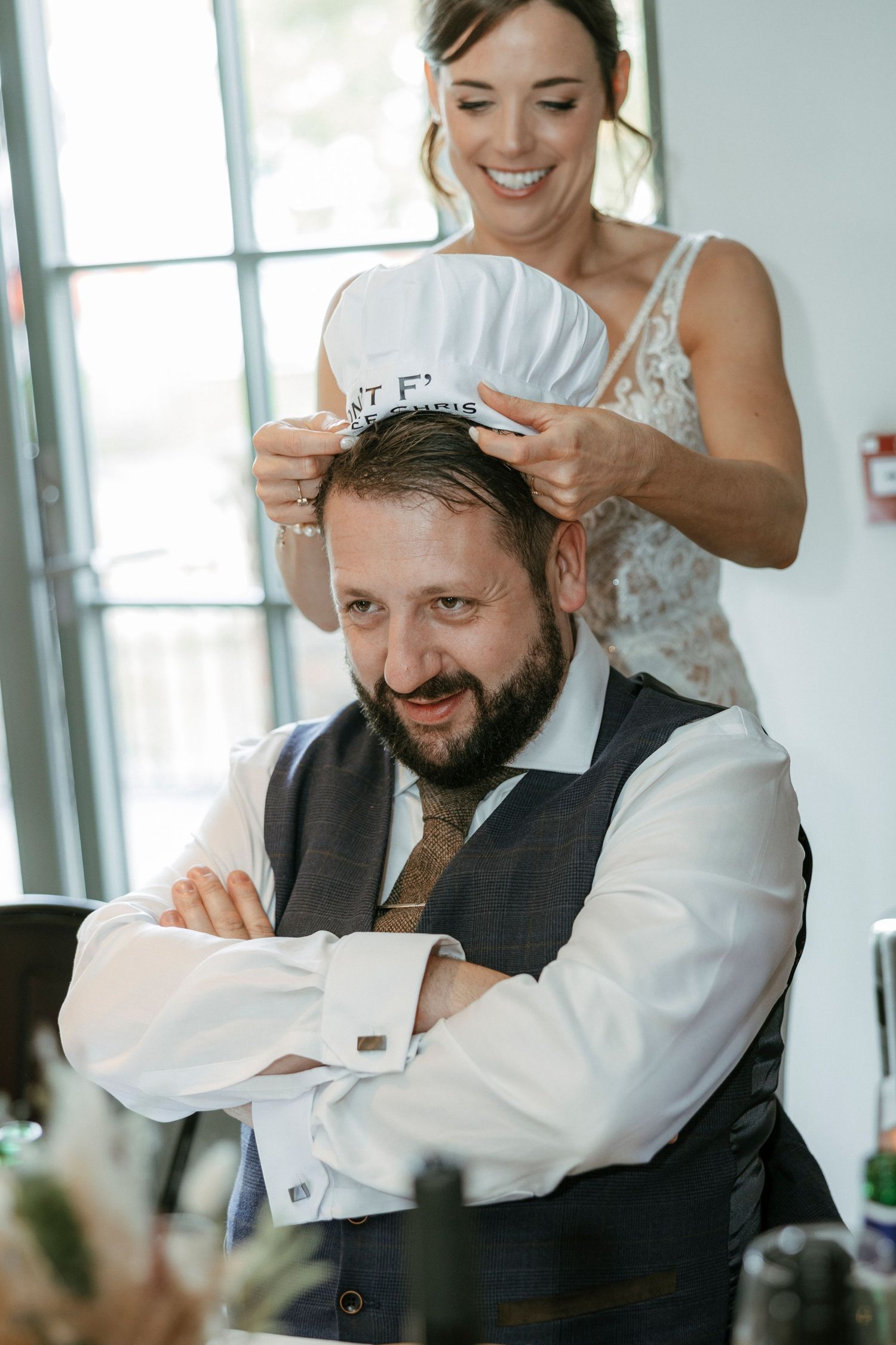 Bride placing a white cap on groom's head at wedding reception, both smiling joyfully.
