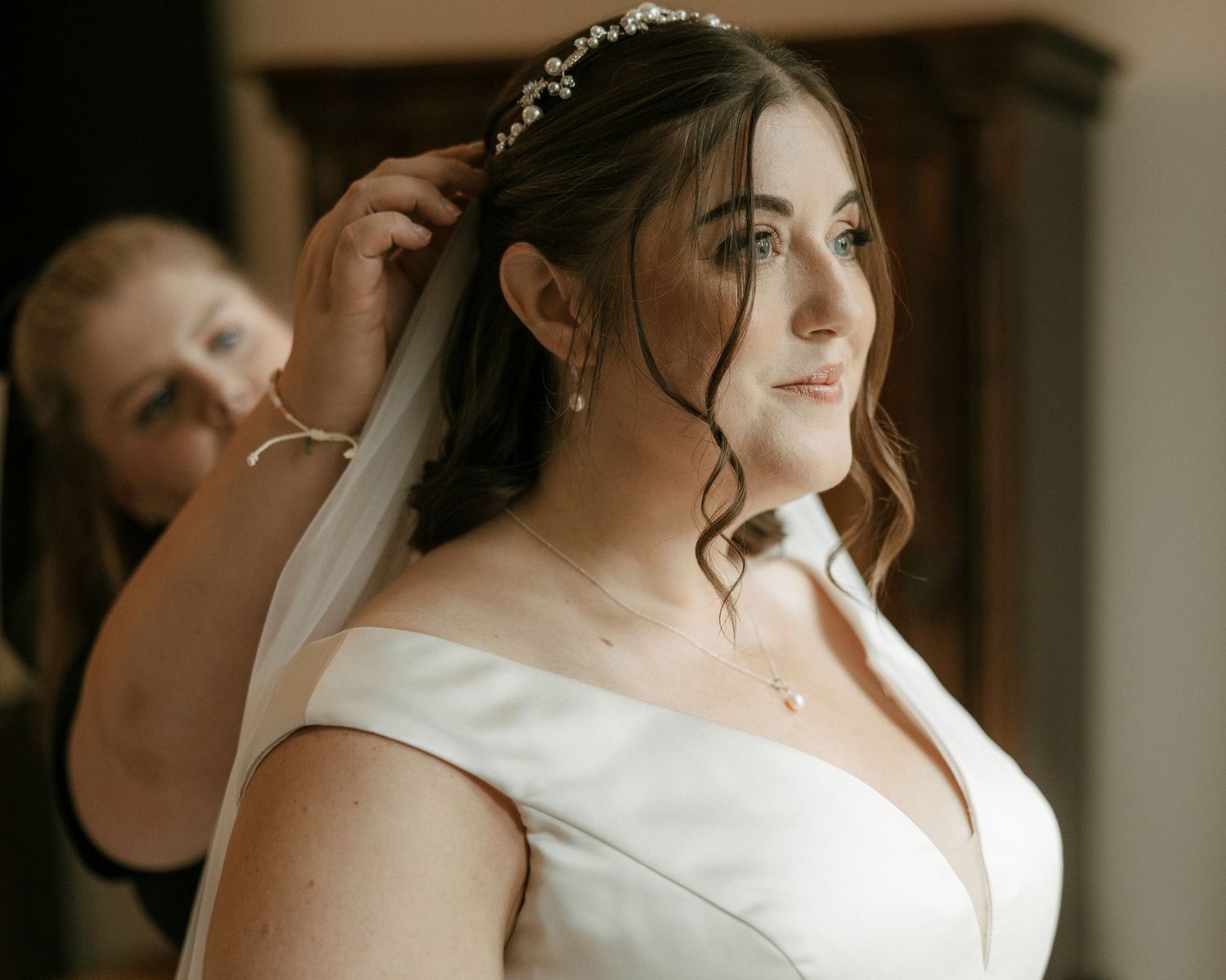 Bride in white gown getting veil placed by bridesmaid, wearing pearl earrings and floral hair accessory.
