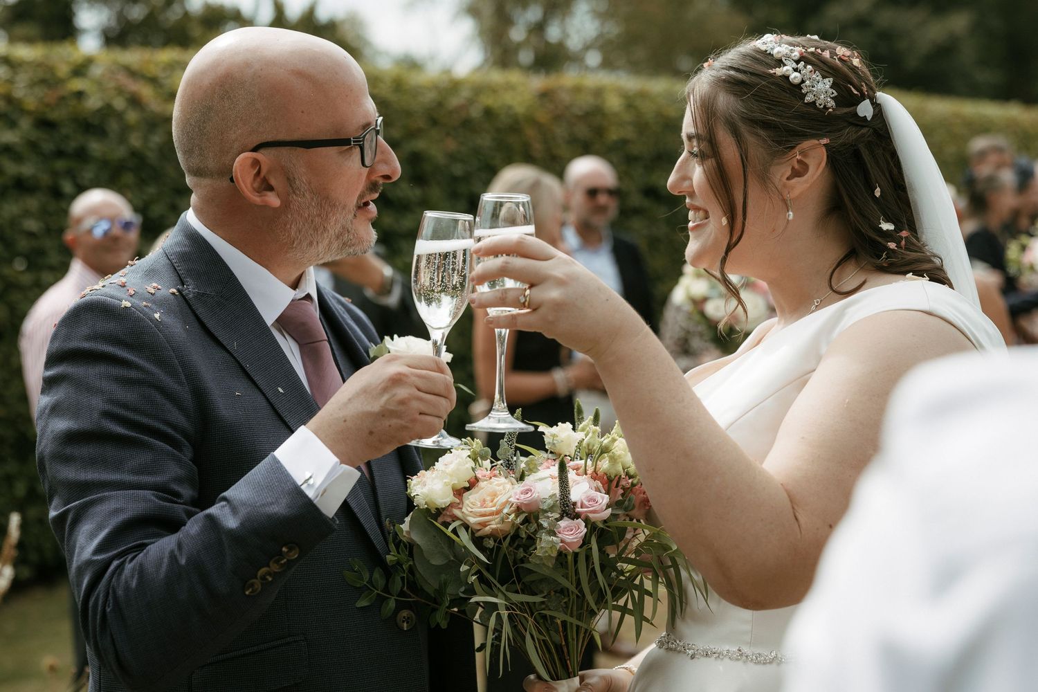 Bride and groom toasting with champagne glasses outdoors after wedding ceremony holding floral bouquet.