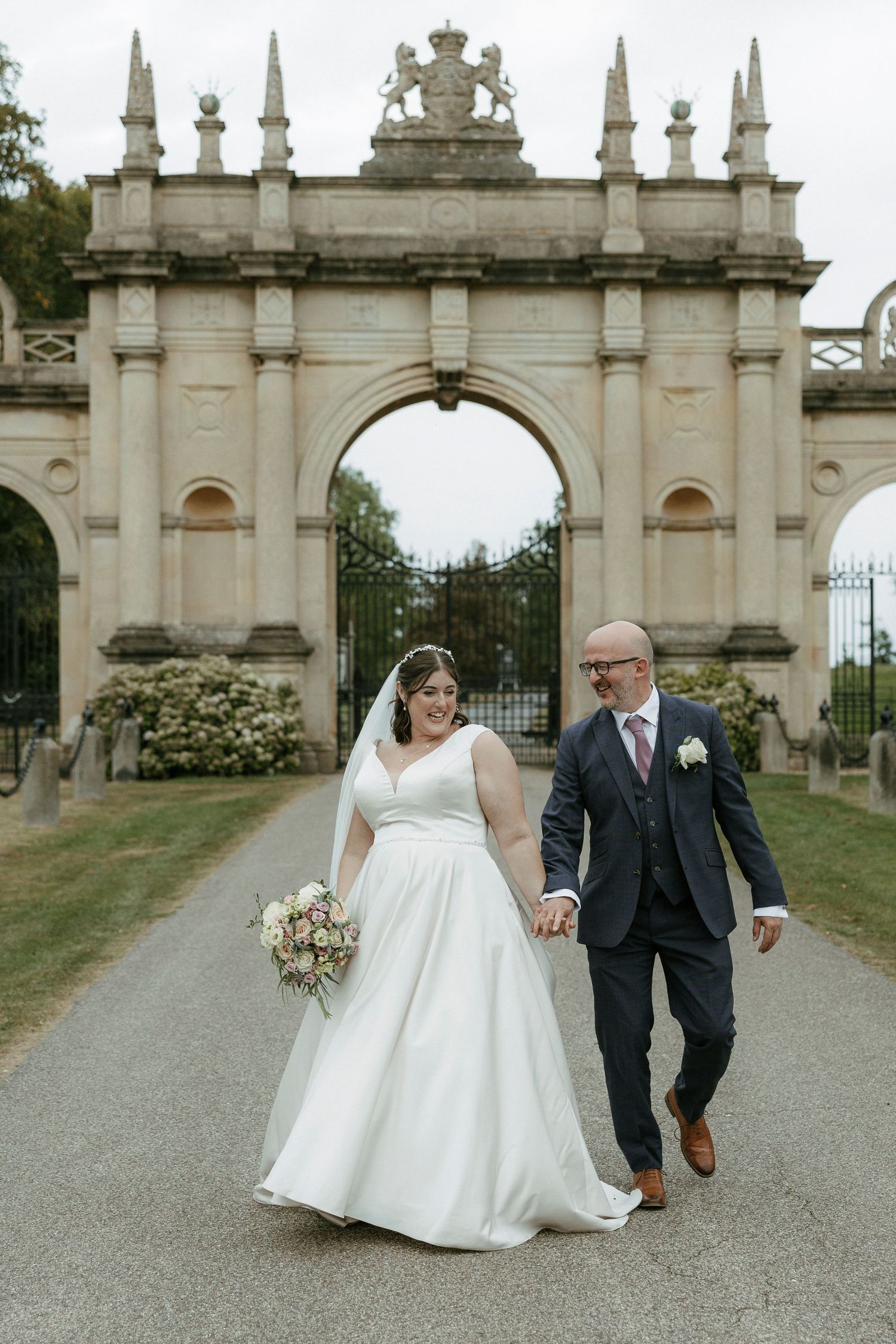 Bride and groom walk hand in hand before an ornate stone archway gate.